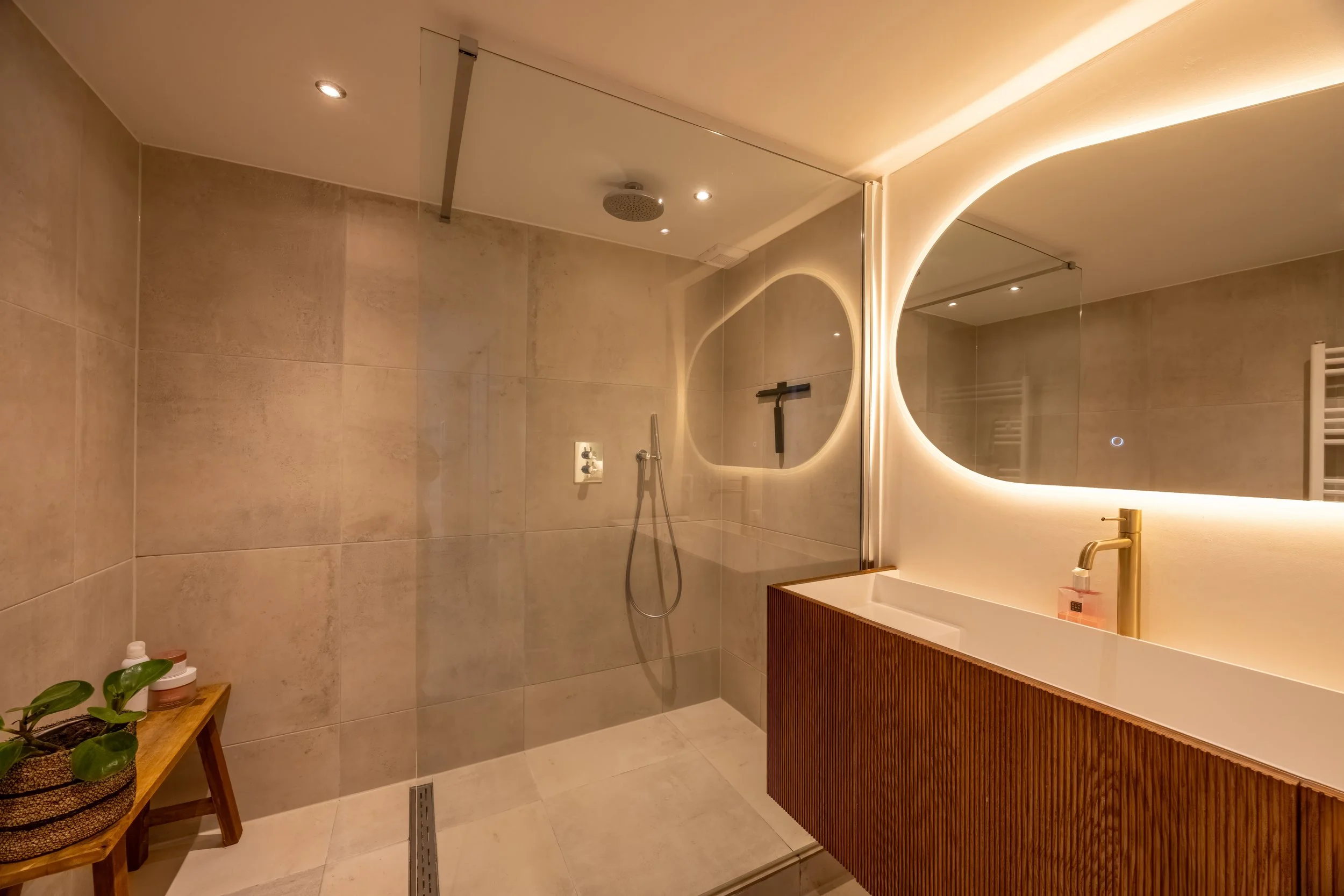 Moderne badkamer met grote spiegel, houten kast, wastafel, inloopdouche, verlichting en beige tegels.