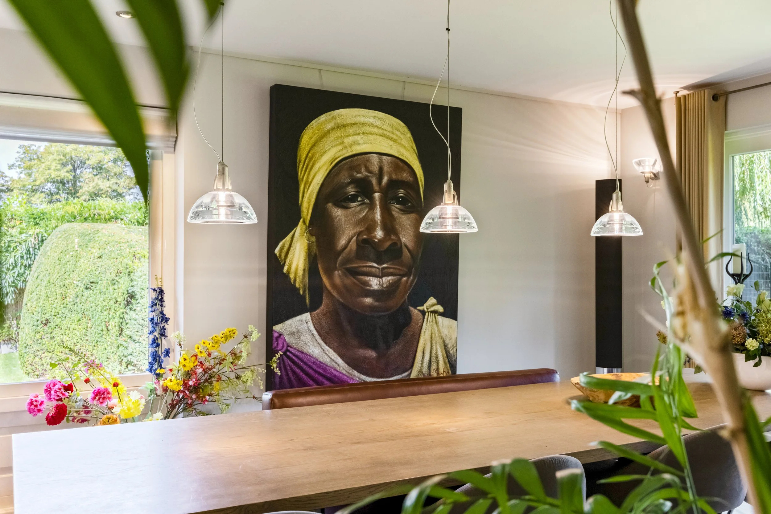 Interieur van een eetkamer met een houtentafel, bloemen, portret van een vrouw met een gele doek op haar hoofd, ramen met uitzicht op een groene tuin, hangende lampen, bloemen en planten.