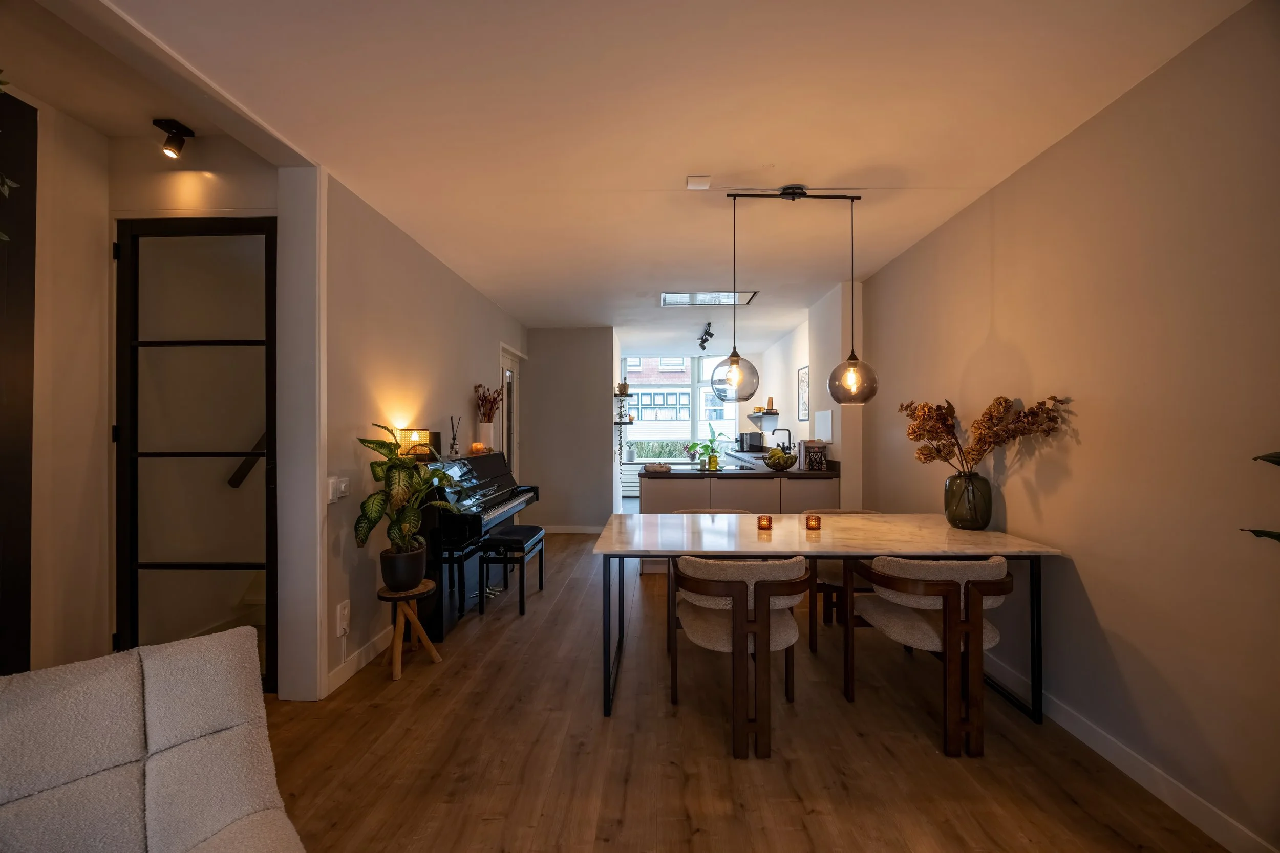 Modern open-plan living en eetkamer met houten vloer, een eettafel met vier stoelen, een keuken in de achtergrond, hangende gloeilampen, een piano met planten en sfeerverlichting.