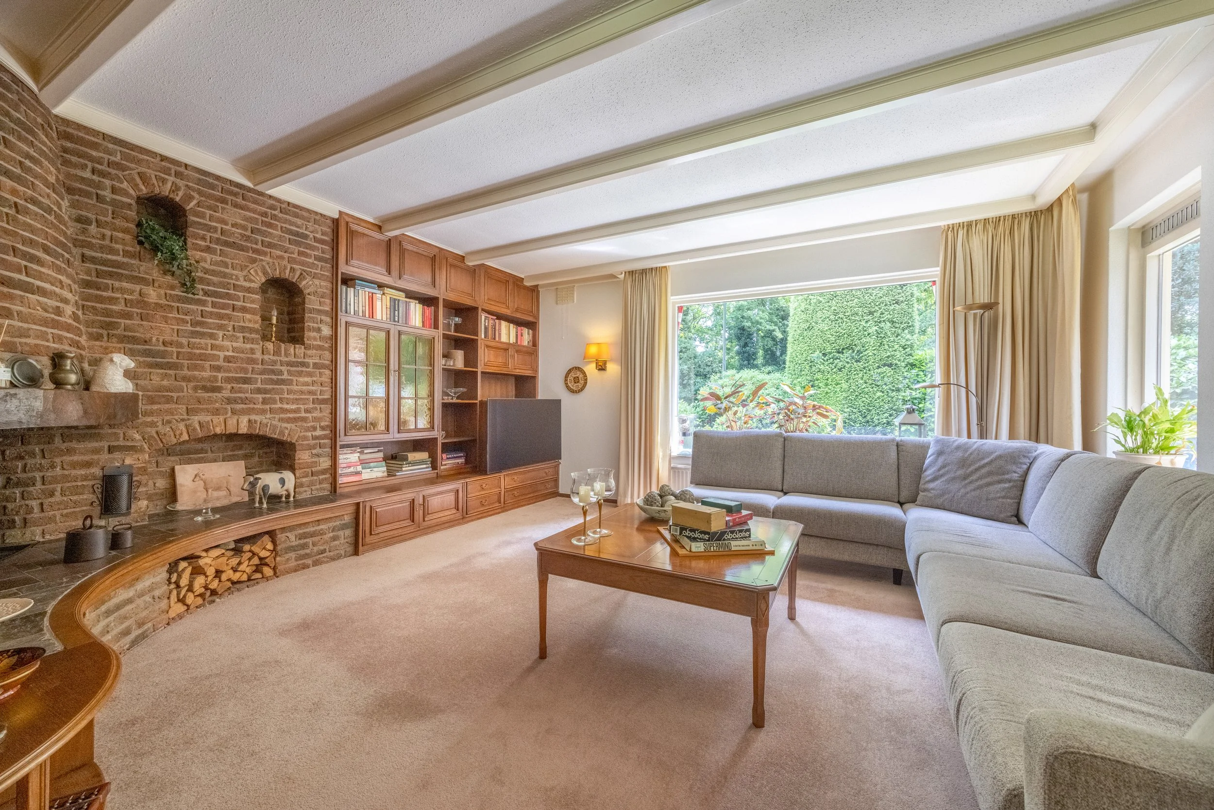 Gezellige woonkamer met een grote hoekbank, een houten salontafel, een ingebouwd boekenrek met tv, een open haard met hout, en grote ramen met uitzicht op groene tuin.