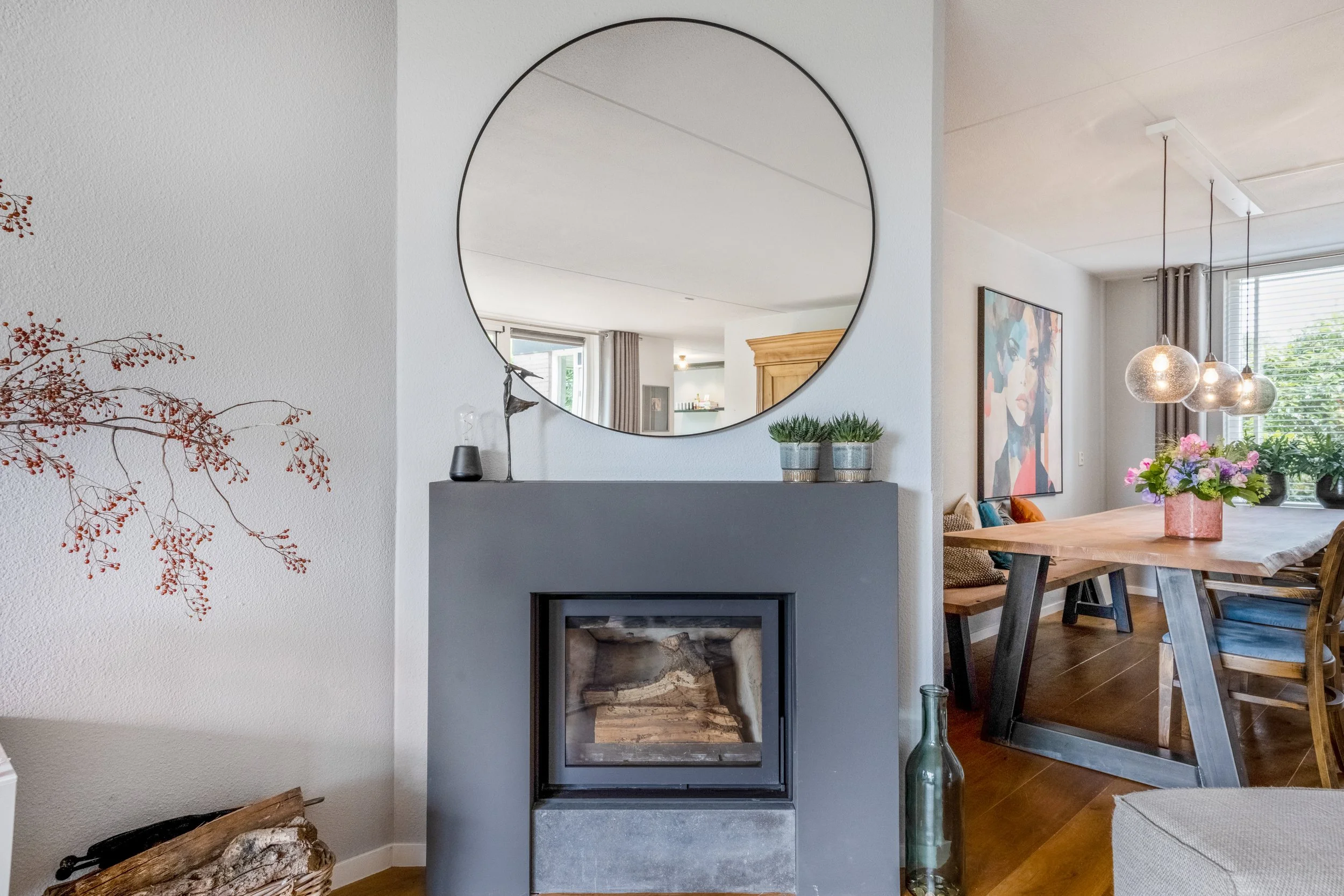 Interieur van een woonkamer met een moderne haard, een ronde spiegel erboven, een houten tafel met bloemen, en een schilderij aan de muur.
