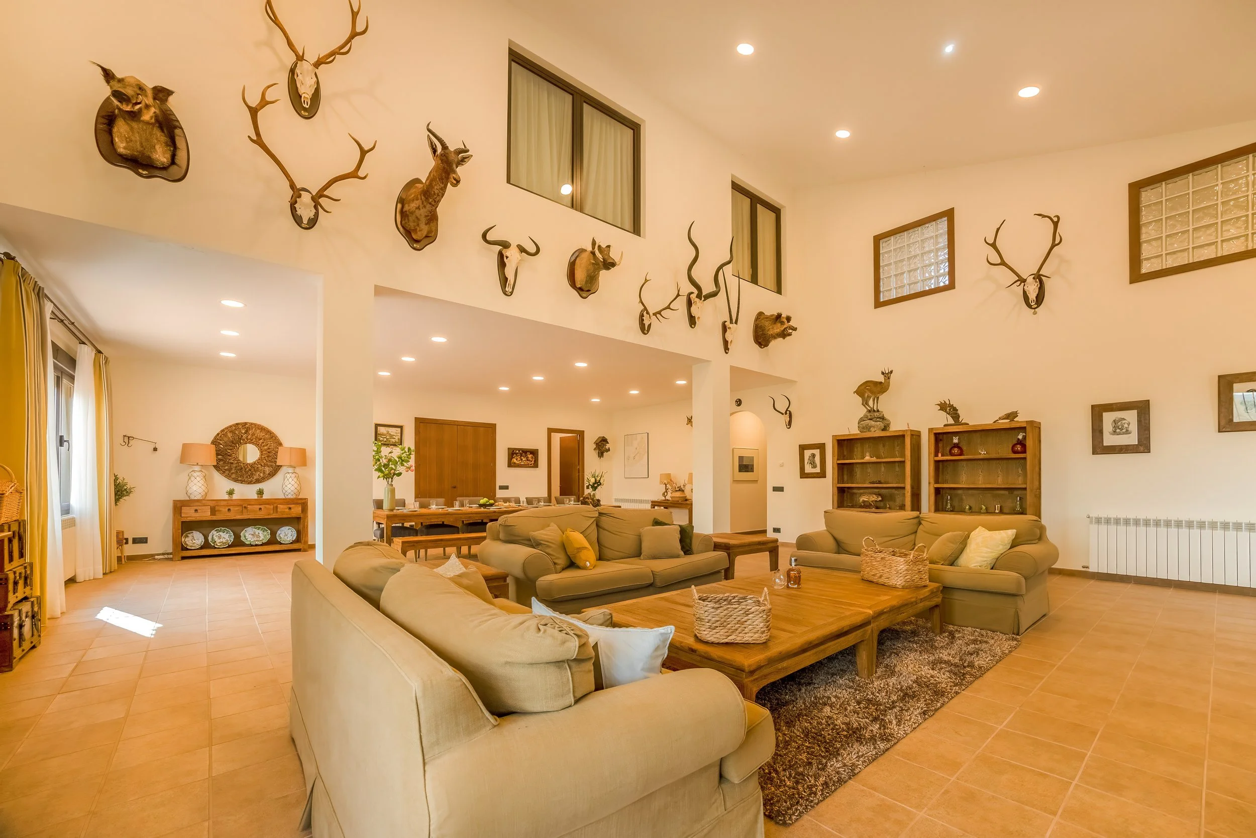 Woonkamer met beige banken, houten salontafel, wanddecoraties van geweien, schilderijen en beeldhouwwerken, keramieken potten, grote ramen met gordijnen, houten meubels en een vloer met terracotta tegels.