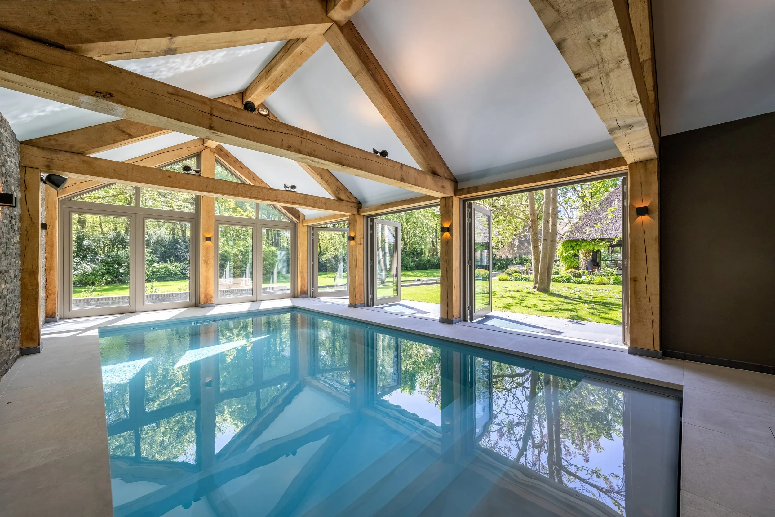 Binnenzwembad met grote ramen die uitzicht bieden op een groene tuin, houten balken en een spiegelend wateroppervlak.