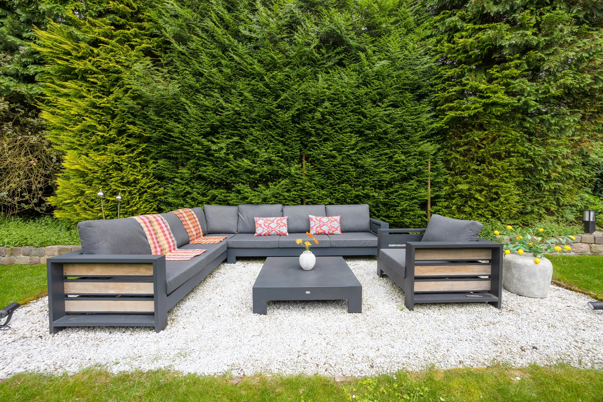 Een buitenbank met grijze kussens en kleurrijke kussens, een rechthoekige salontafel met een vaas met gele bloemen, twee lampen, een grote pot met citroenplanten, allemaal op een grindoppervlak voor een hoge groene heg.