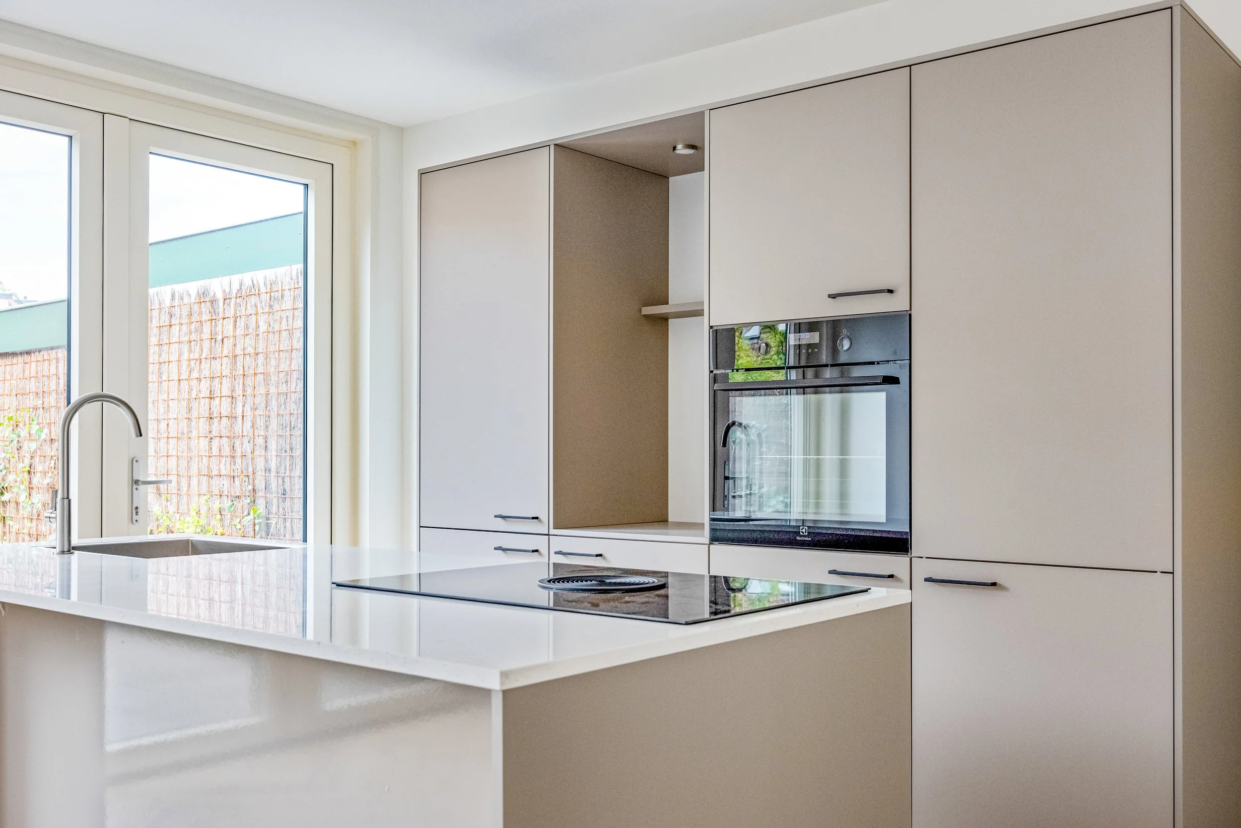 Moderne keuken met witte kasten, inbouwoven, inductiekookplaat en een keukenaanrecht met een gootsteen, grote ramen naar buiten. 