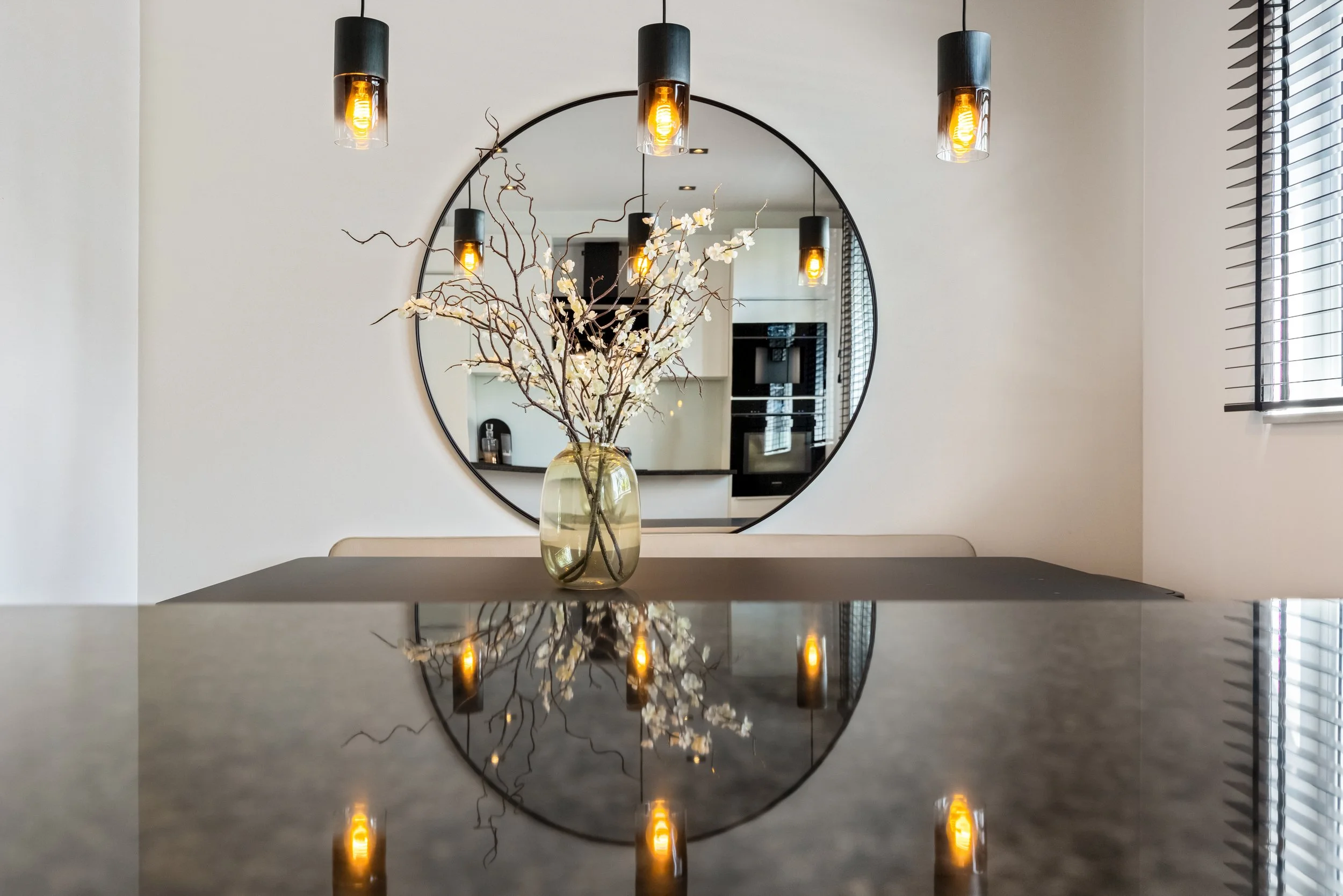 Interieur van een modern huis met een tafel, een vaas met witte bloemen, een ronde spiegel en vier hanglampen.