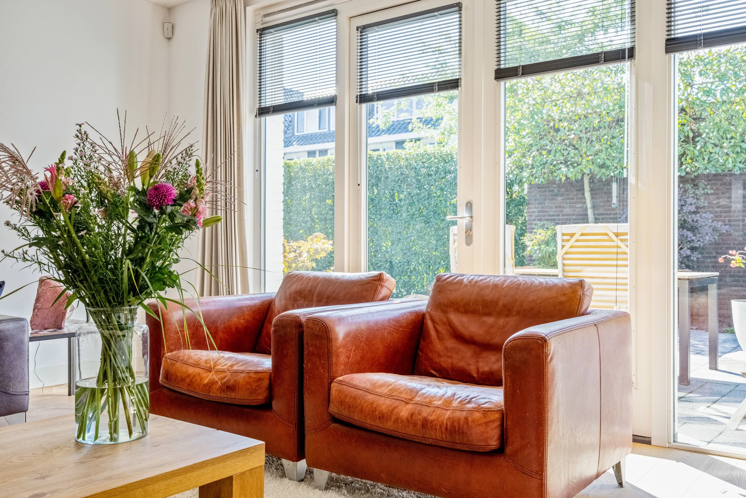 Binnenkamer met twee oranje leren stoelen, een grote vaas met bloemen op een houten tafel, grote ramen met horren en uitzicht op een tuin.