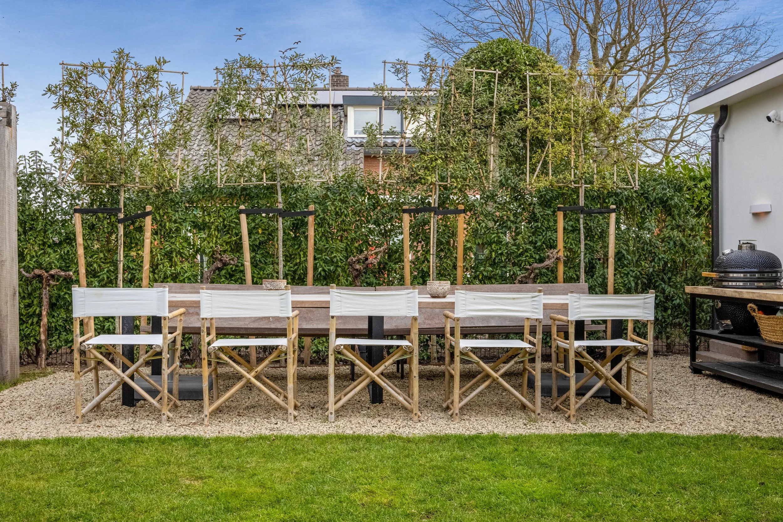 Tuin met tapas tafel en zes klapstoelen, omgeven door groene struiken en boomtoppen.
