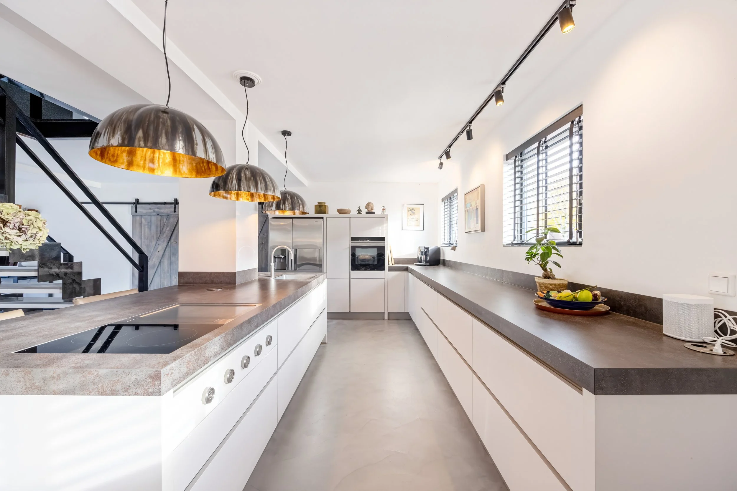 Moderne keuken met witte kasten, een grijze countertop, drie hanglampen en drie ramen met jaloezieën, decoratieve planten en keukenapparatuur.