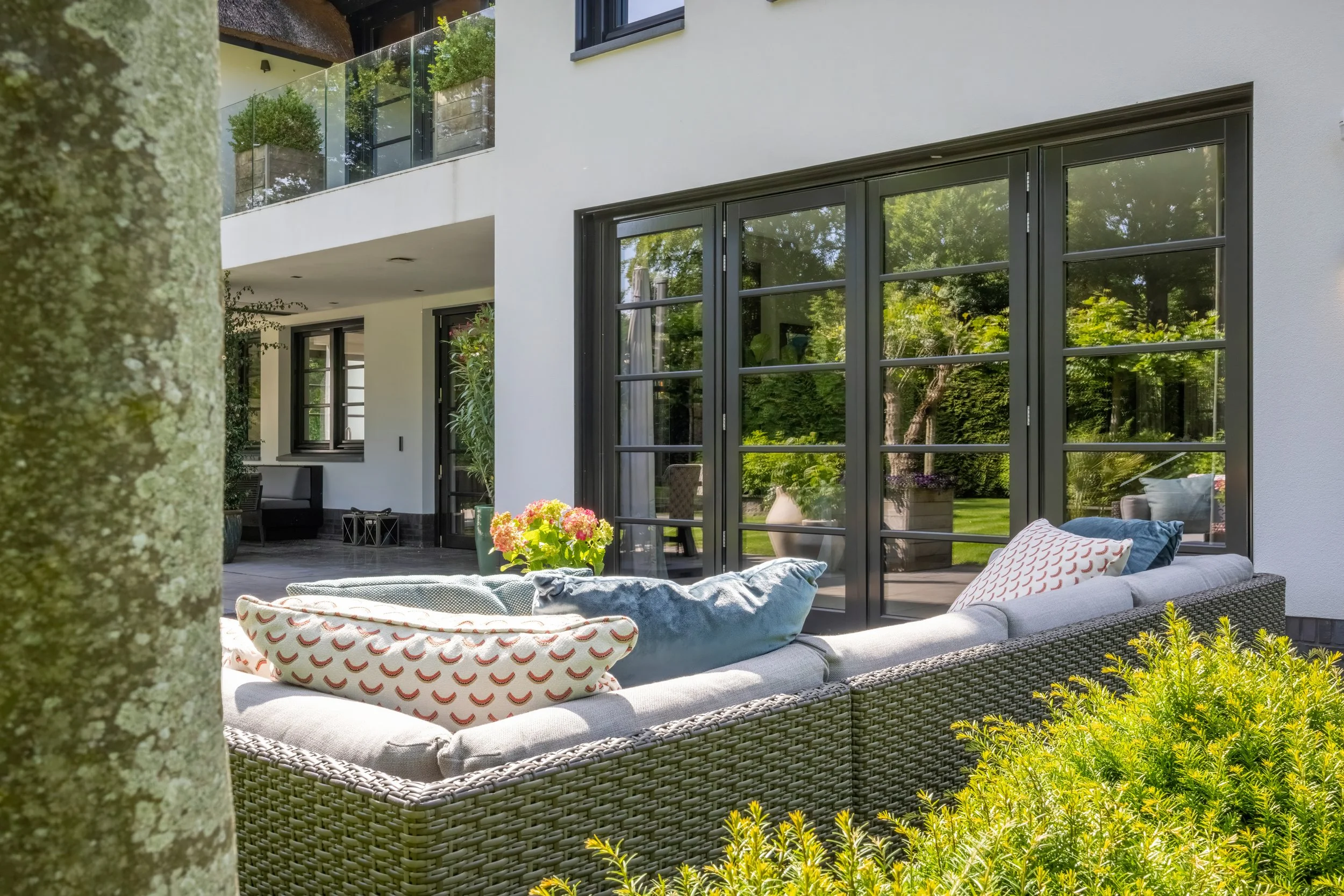 Gezellige patio met wicker bank, kussens, planten en uitzicht op tuin van een modern huis.