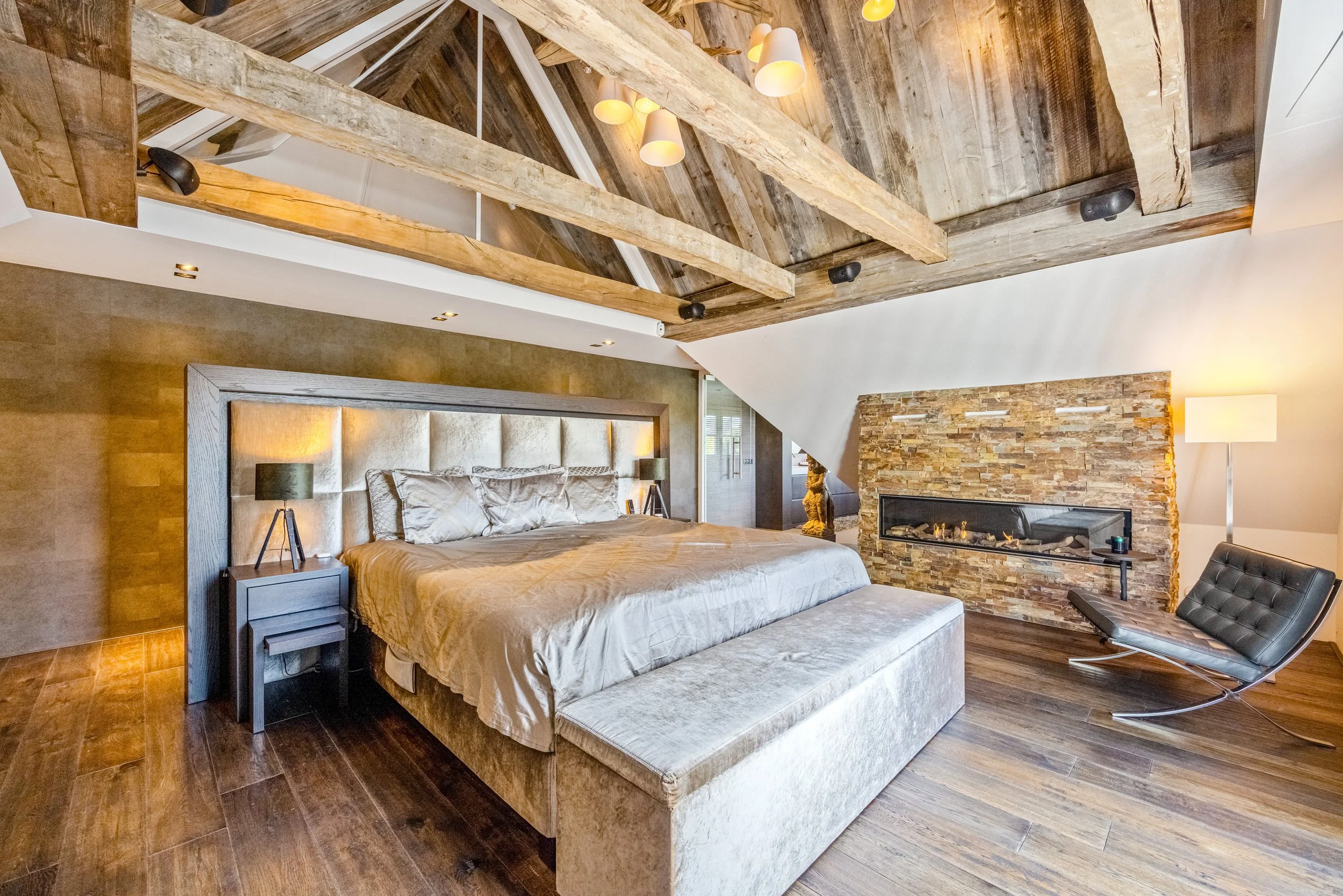 Een moderne slaapkamer met een groot bed, een leren fauteuil, een stenen haard en een plafond met houten balken.