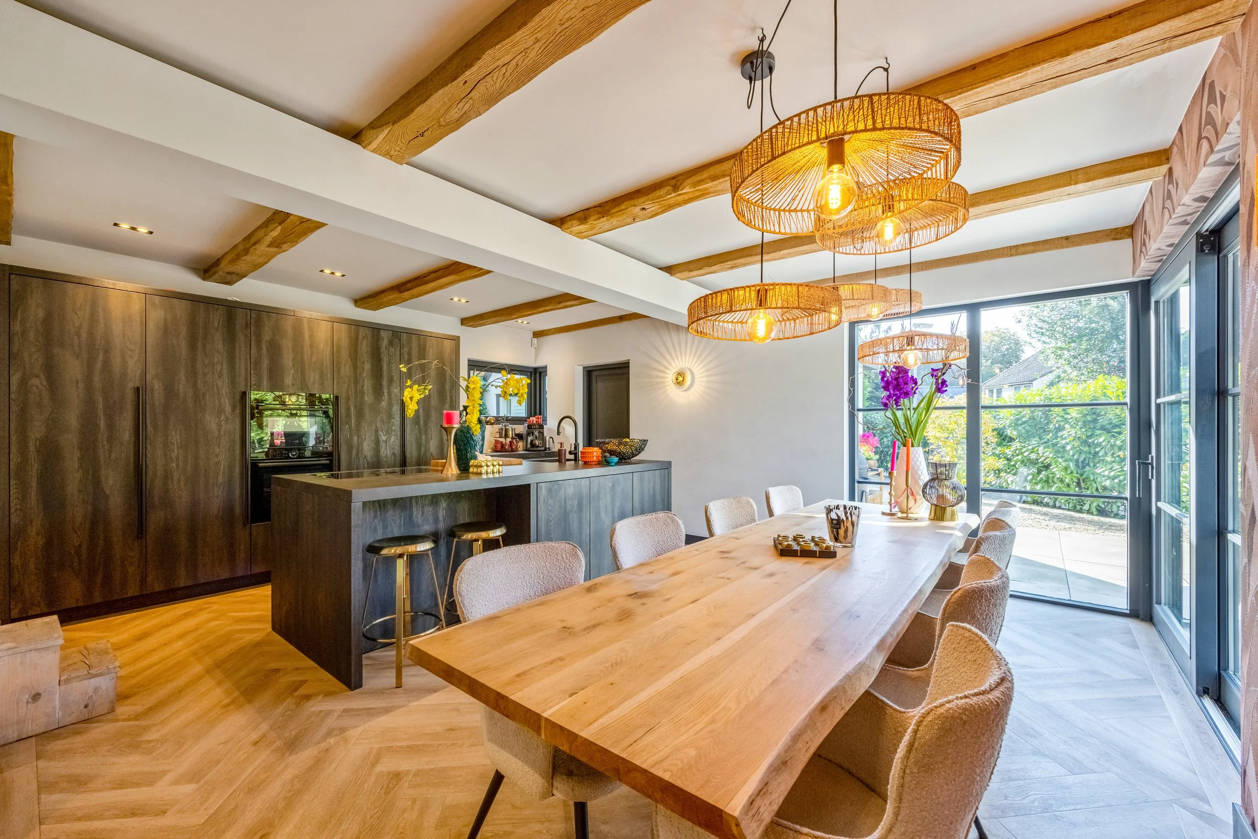 Een modern eetkamer met een grote houten tafel, beige stoelen, een keuken met een donker houten kast en een keukeneiland, grote ramen met uitzicht op een tuin, en decoratieve hanglampen.