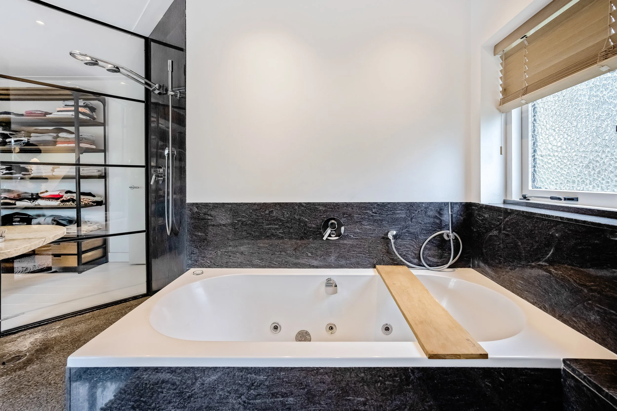 Een moderne badkamer met een inbouw Jacuzzi-bad, omgeven door zwarte marmeren muren, naast een raam met houten jaloezieën.