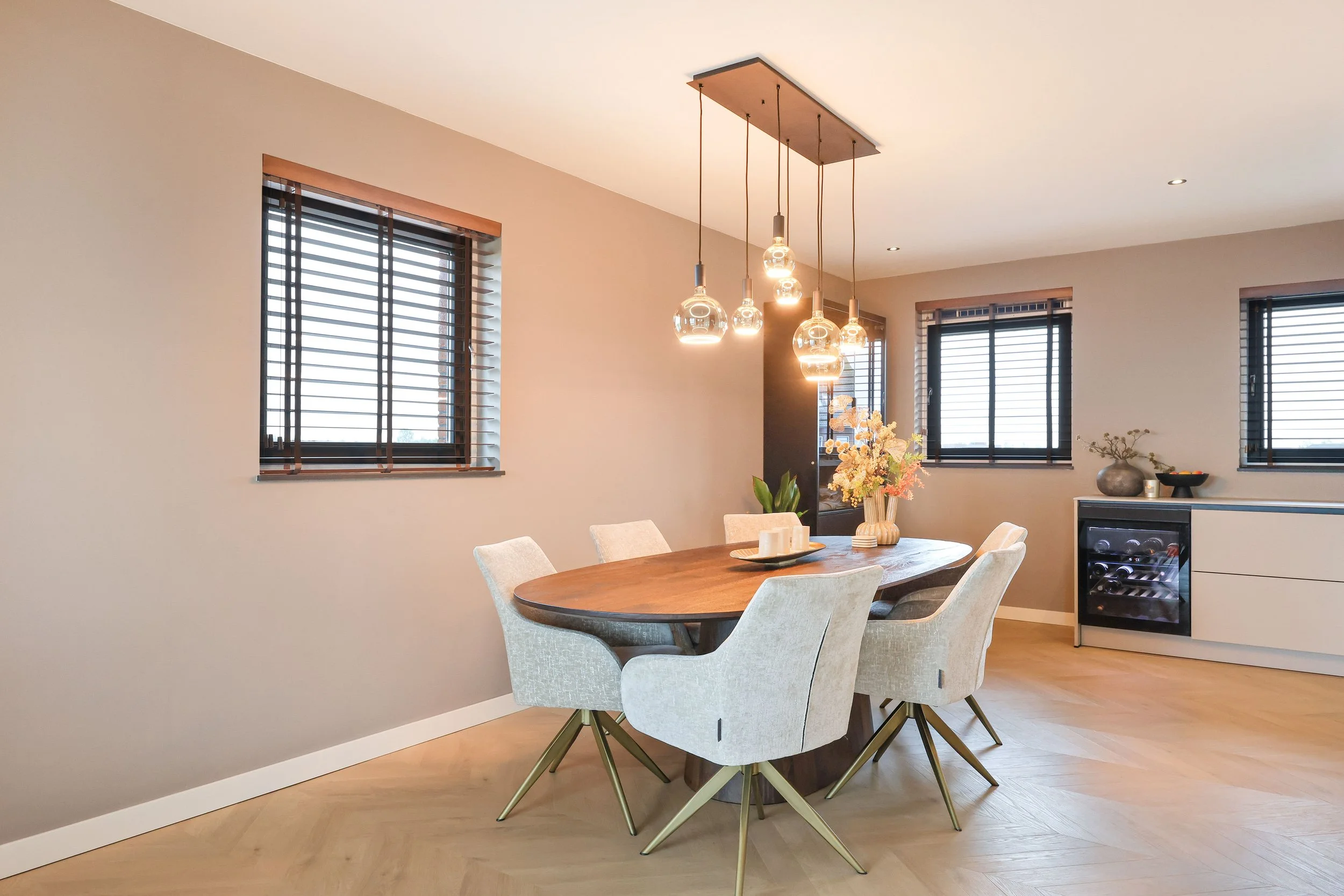 Eethoek met een ronde houten tafel, zes lichtgrijze stoelen en een bloemenvaas, met een pendelverlichting en drie ramen met houten jaloezieën.