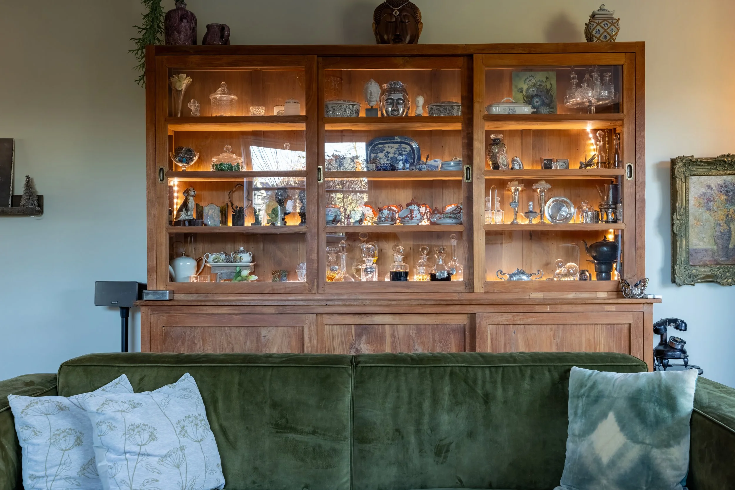 Een grote houten vitrinekast gevuld met kristallen en decoratieve voorwerpen, geplaatst boven een groene bank met witte en groene kussens in een woonkamer.