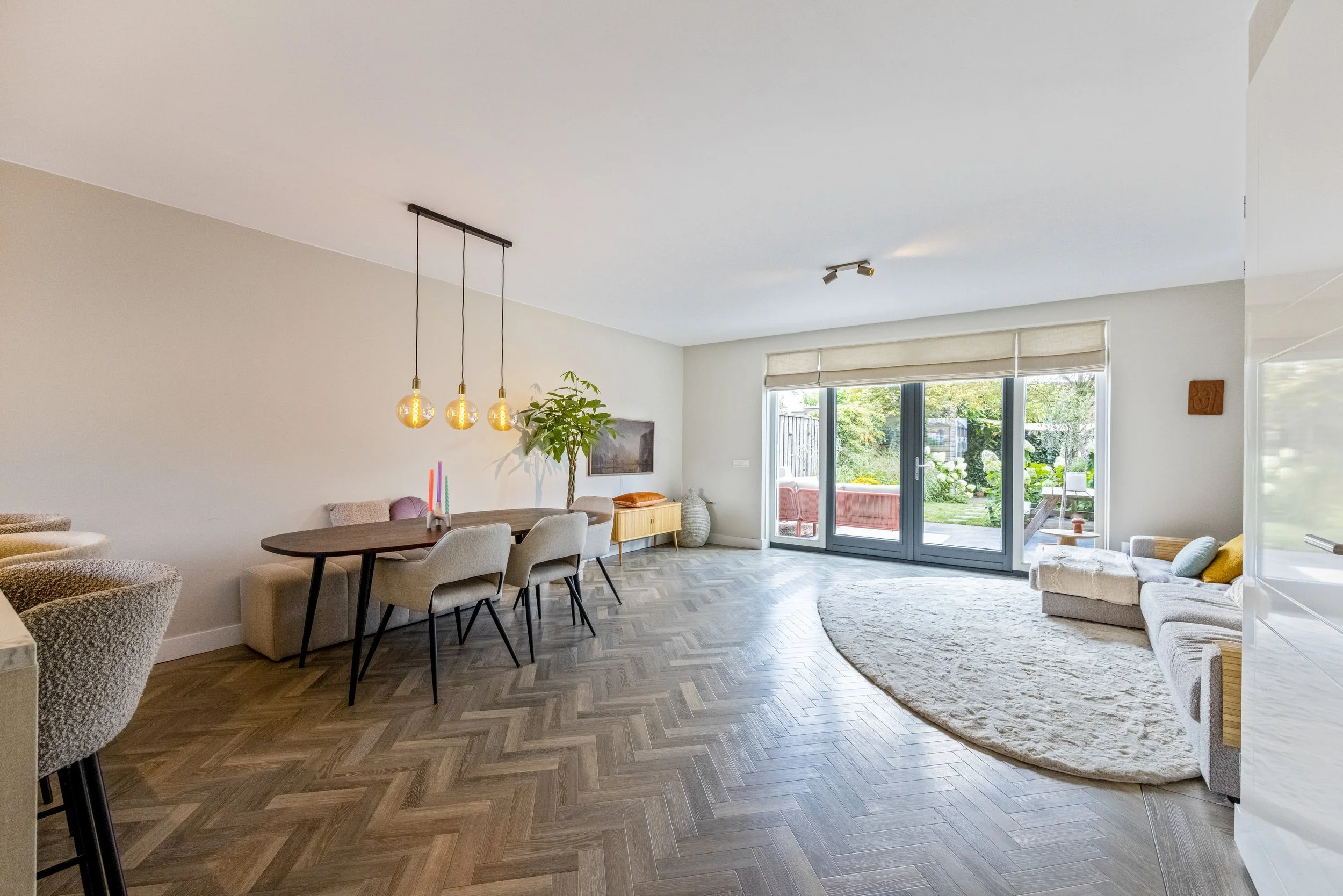 Woonkamer met grote glazen schuifpui naar tuin, eettafel met zes stoelen, zithoek met bank en tapijt, houten vloer en planten.