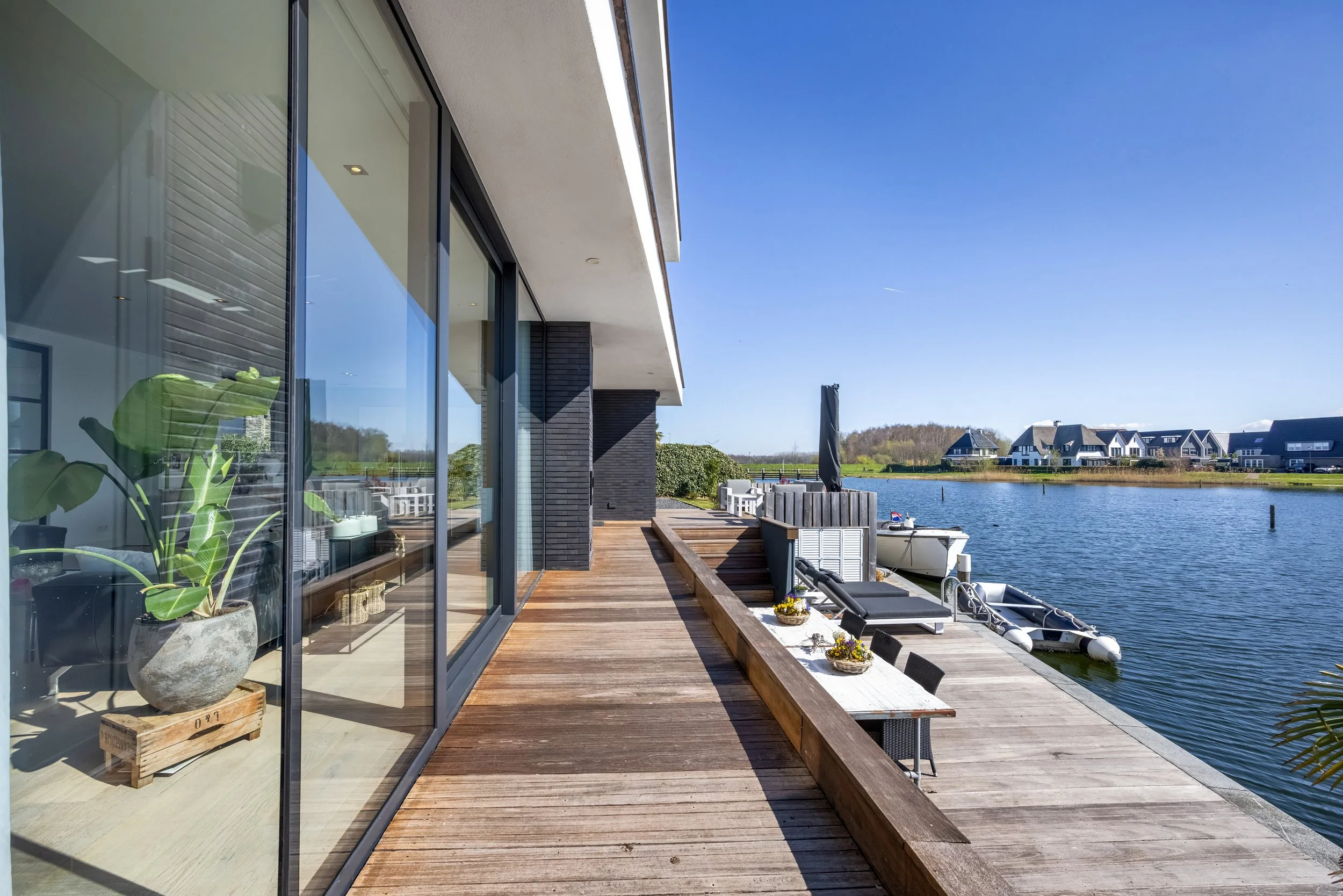 Een moderne woning met een glazen gevel en een houten veranda, uitkijkend over een rivier met boten en huizen in de achtergrond op een heldere dag.