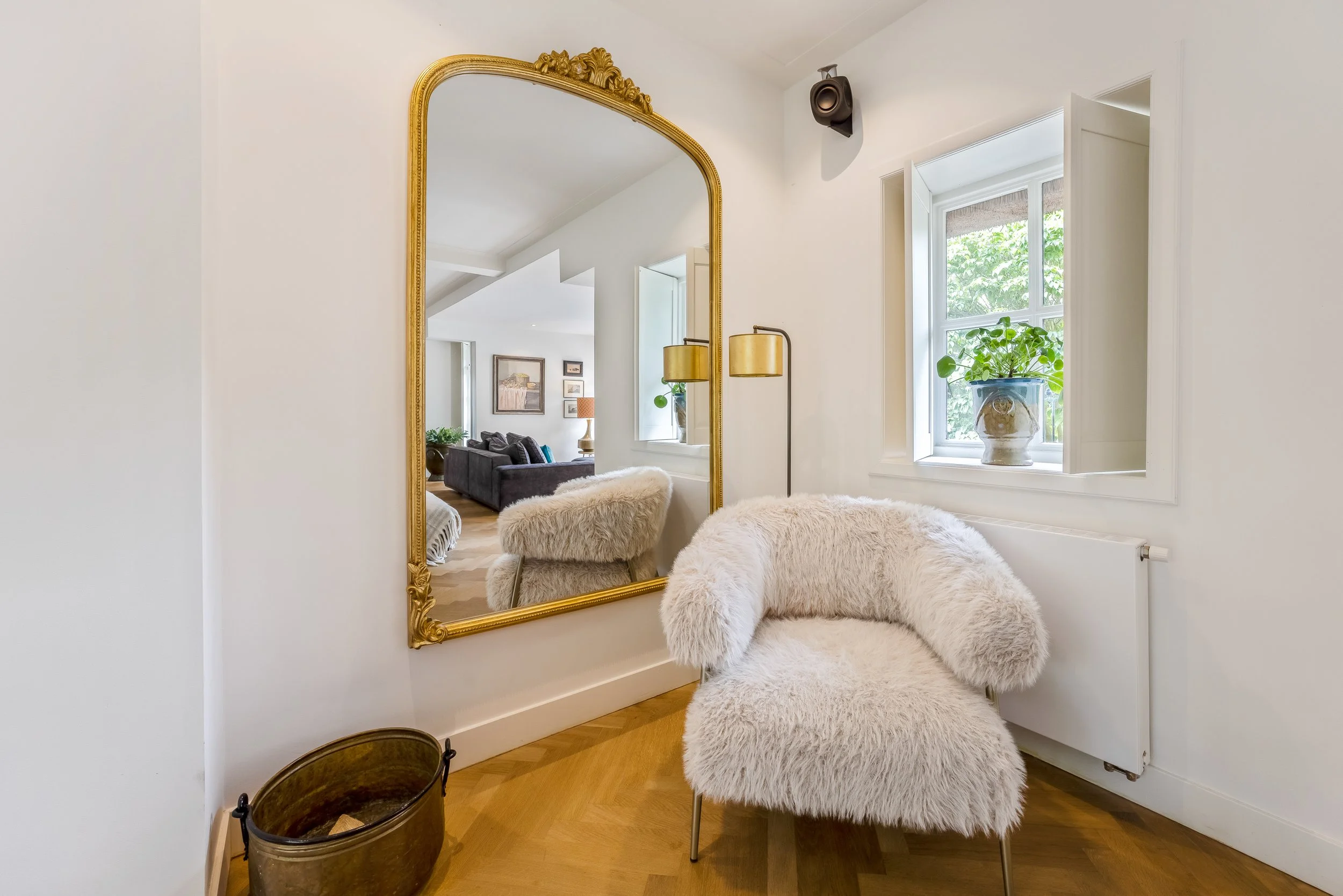Een witte kamer met een grote, gouden spiegel aan de muur, een beige bontkussenfauteuil, een loterlamp, een raam met een plantenbak, en een houten vloer.