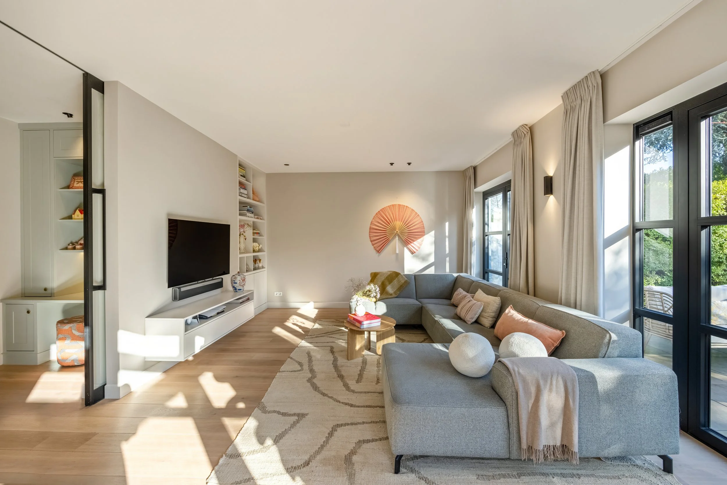 Licht ingericht woonkamer met grote L-vormige bank, houten salontafel, tv aan de muur, ramen met gordijnen, en decoratieve elementen zoals vazen en kussens.