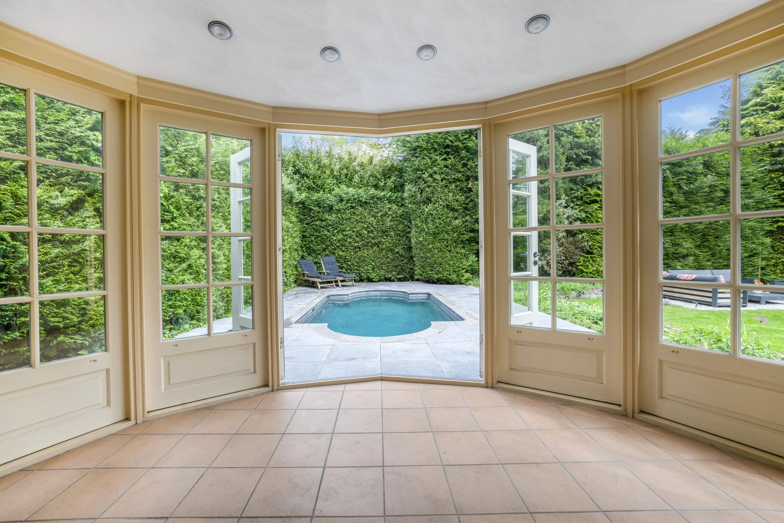 Binnenruimte met grote ramen die uitkijken op een tuin met een zwembad en ligstoelen.