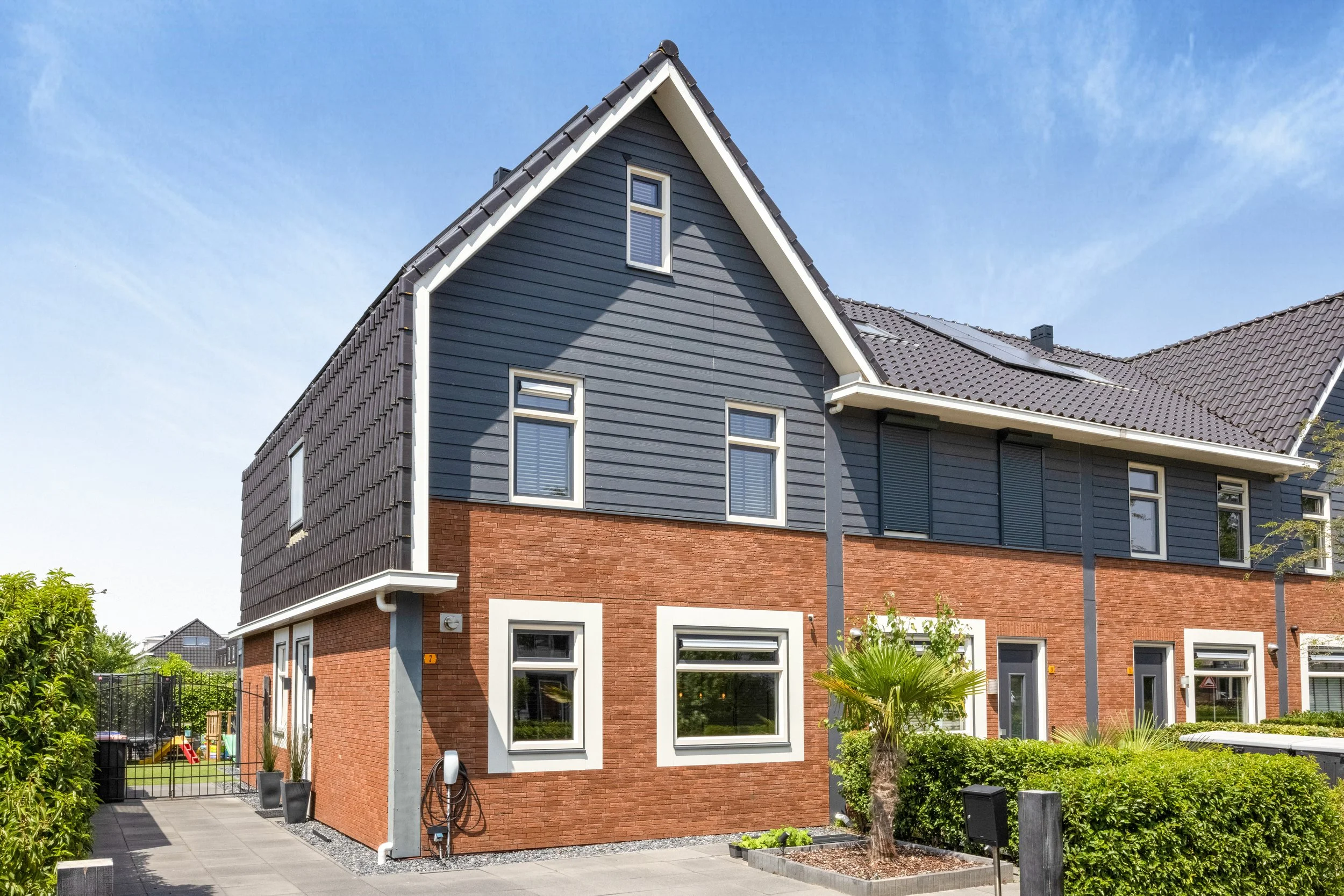 Moderne koopwoning met donkere houten gevel, kleine tuin met palmboom, en een speeltuin in de achtergrond.