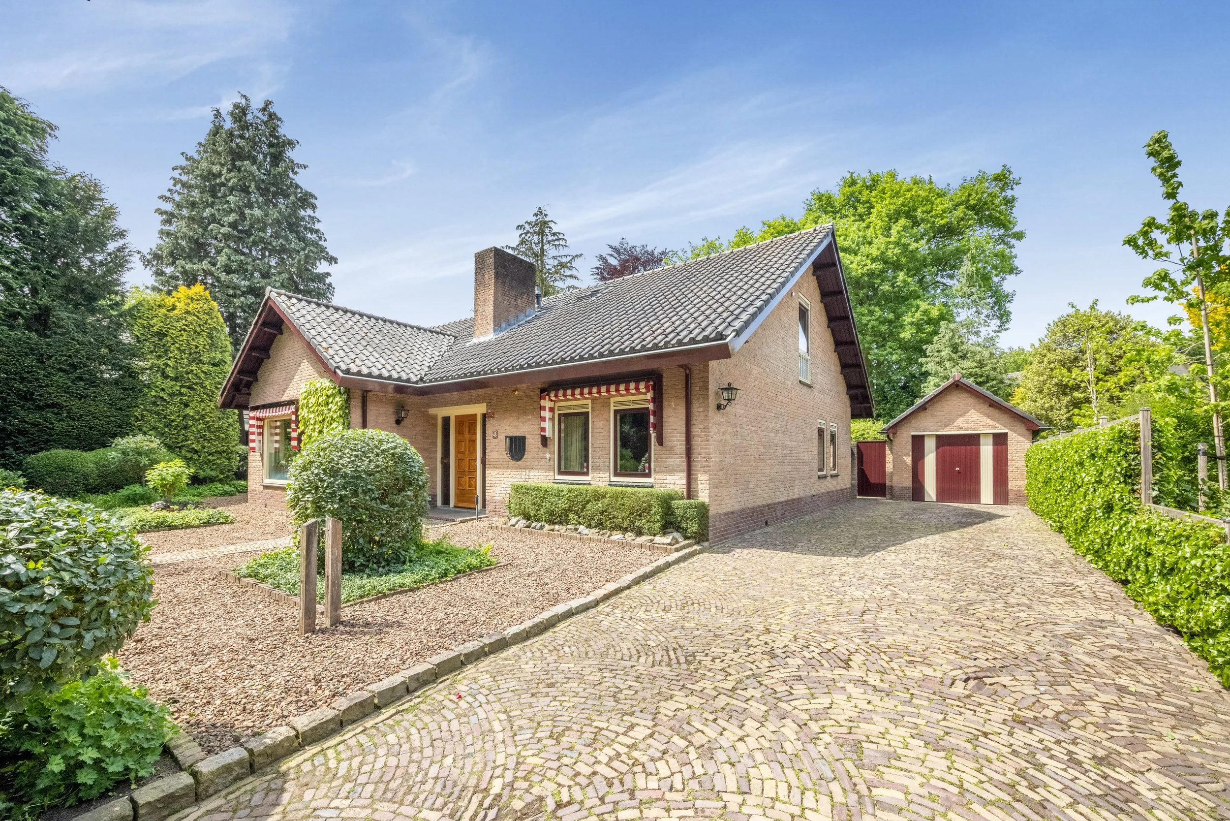 Een huis met een oprit, groene struiken, bomen en een blauwe lucht.