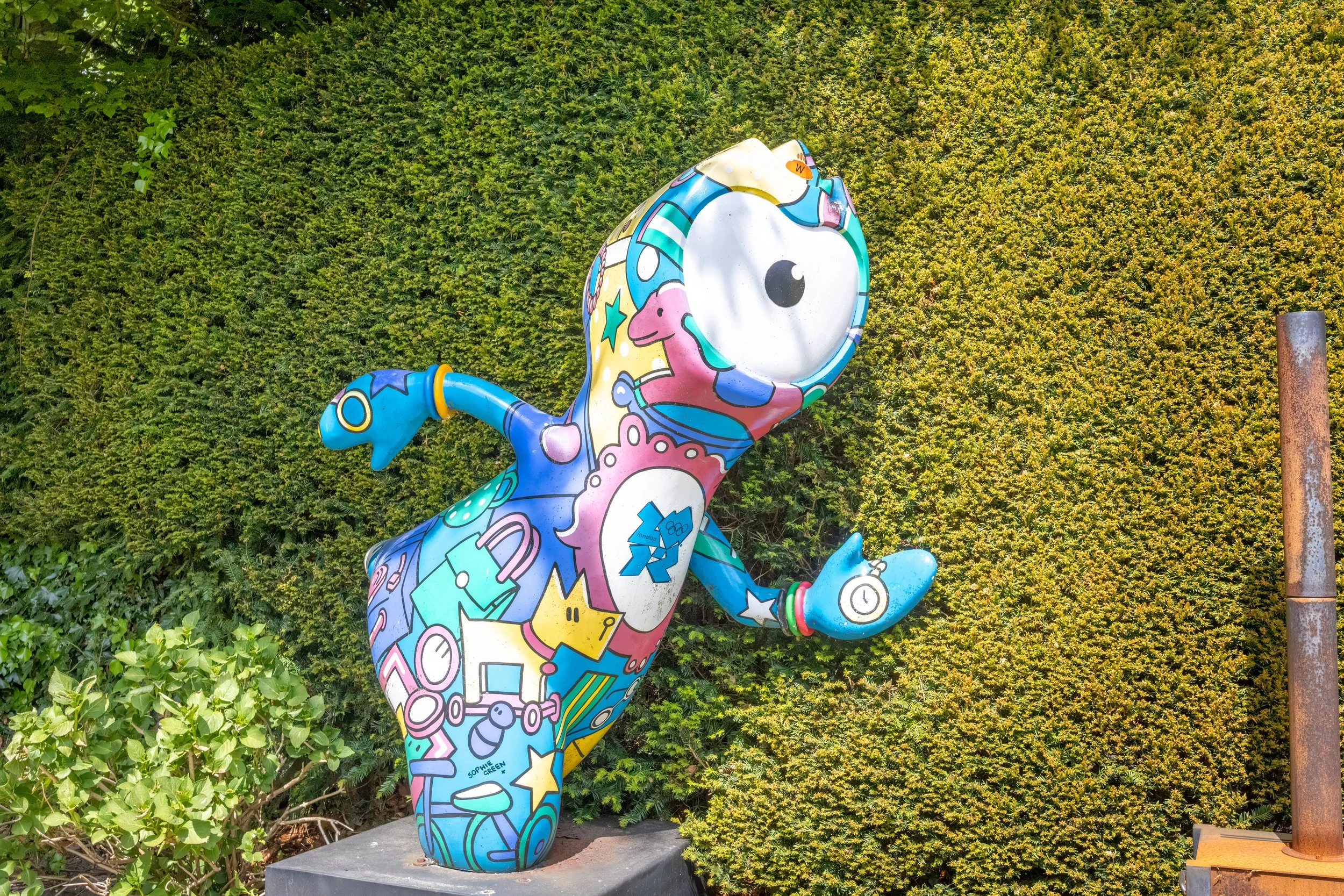 Kleurige straatkunst van een dinosaurus met astronautenthema, uitbundig versierd met symbolen en kleuren, standbeeld op een voetstuk in een tuin of park, met groene heg op achtergrond.