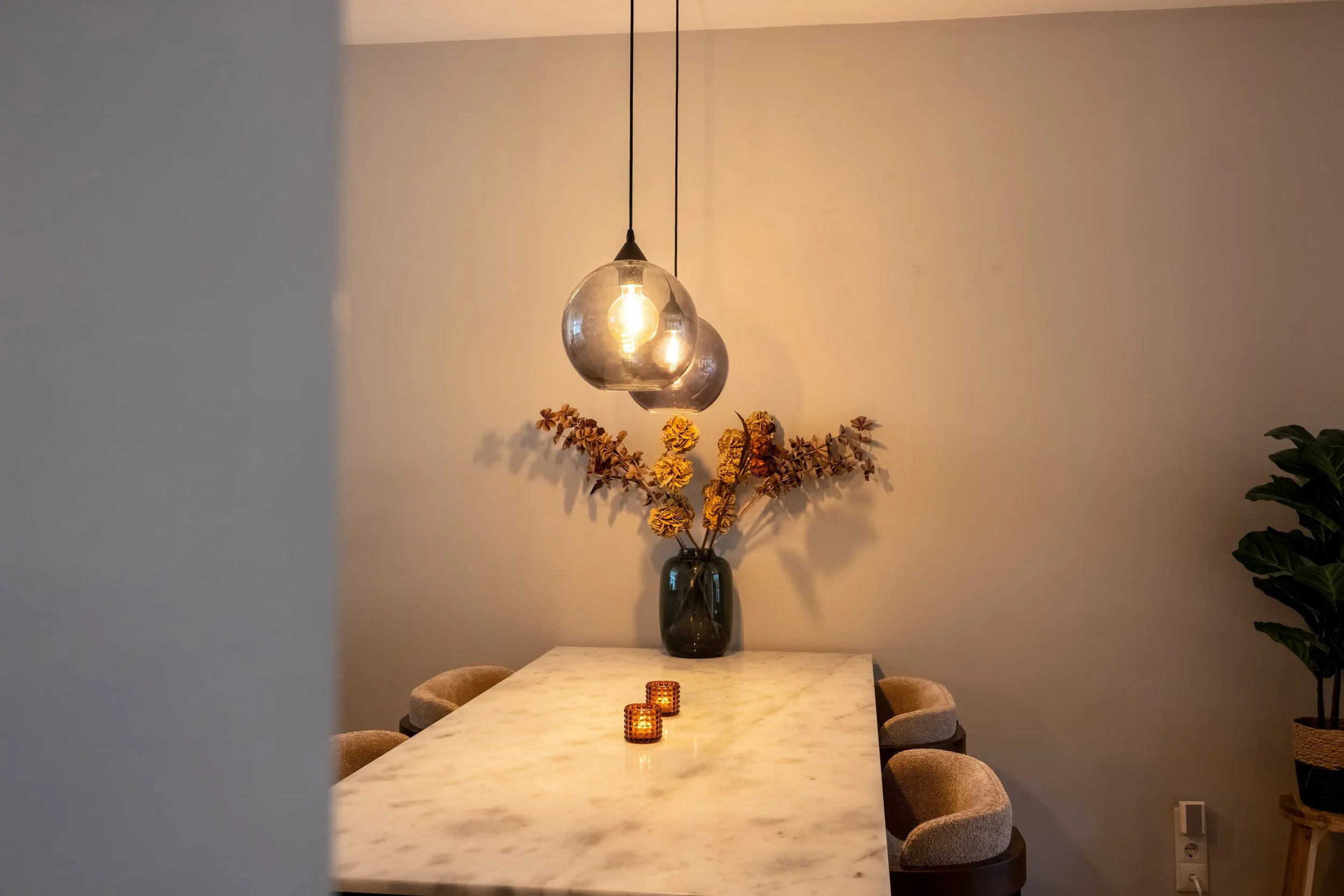 Een eetkamertafel van wit marmer met vijf bijpassende beige gestoffeerde stoelen. Op de tafel liggen twee kleine kaarsen in geurige houders. Aan de muur hangt een donker glazen vaas met gedroogde bloemen en takken. Boven de tafel hangen twee glazen h
