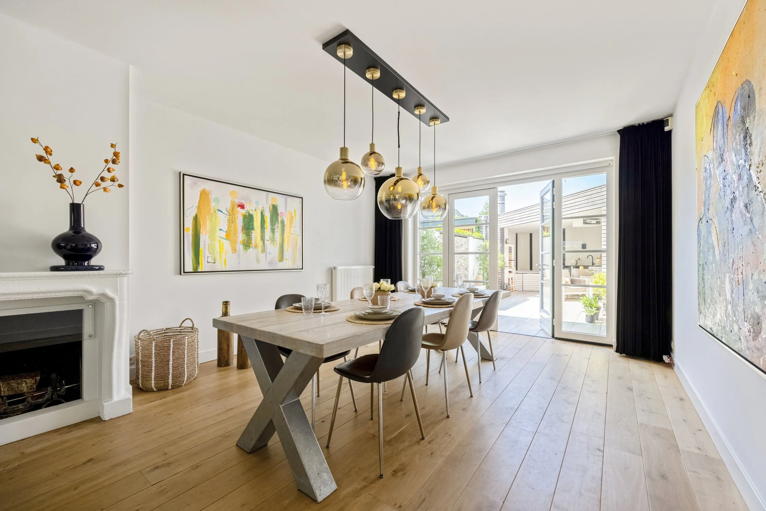Eetkamer met houten tafel, zes stoelen, hangende gouden lampen, schilderijen aan witte muren, open deur naar terras met buitenruimte en zwart gordijn.