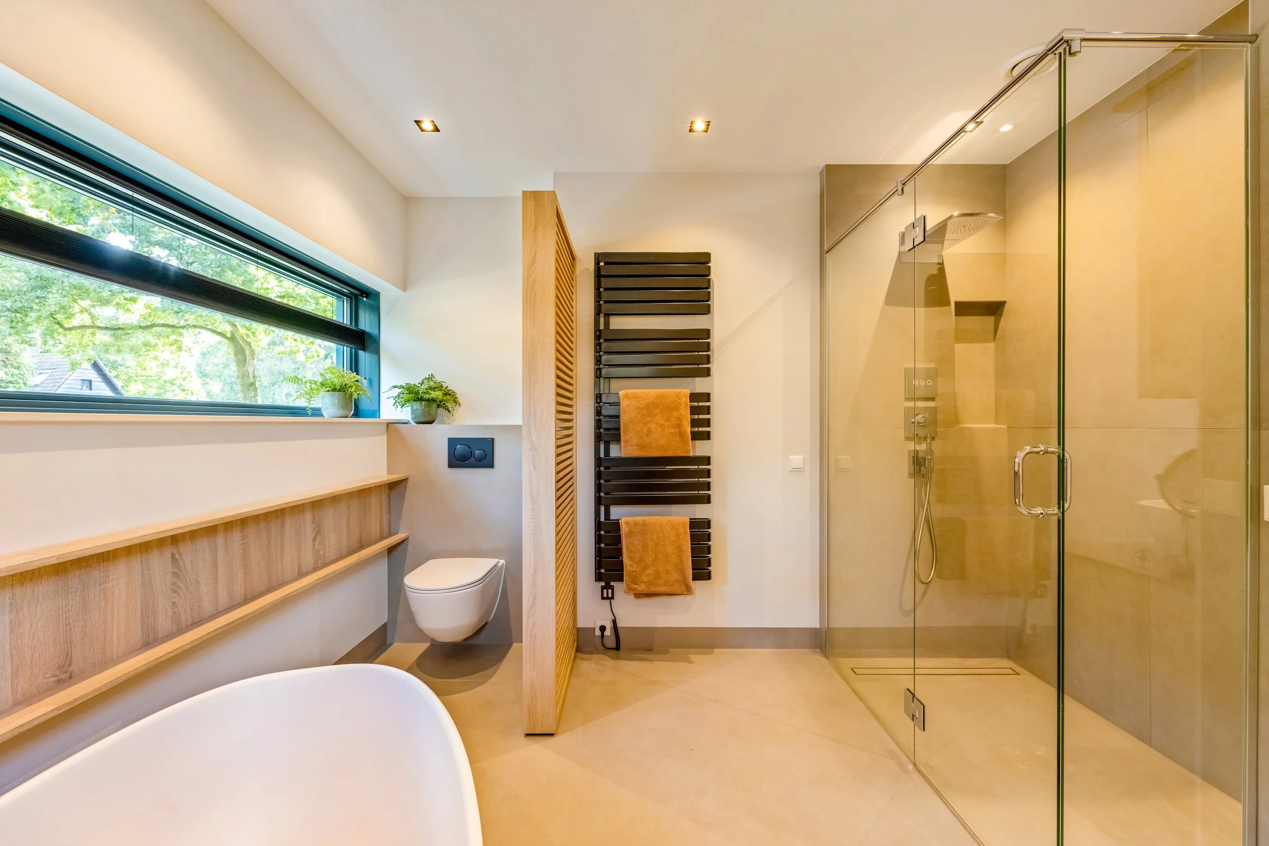 Modern badkamer met douche, toilet en raam met uitzicht op groene bomen, planten op vensterbank.
