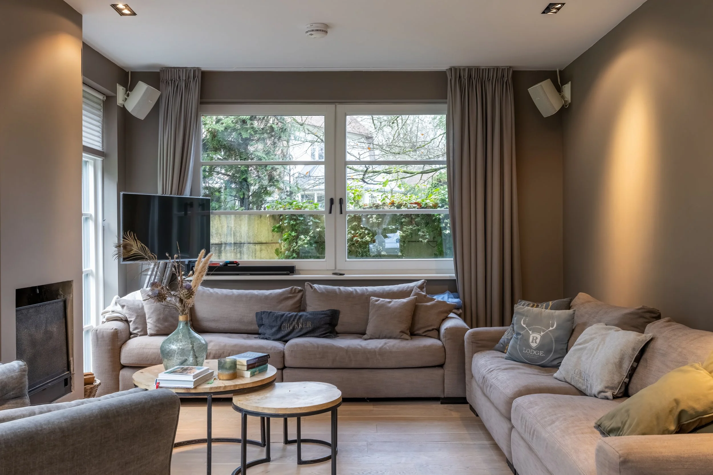 Gezellige woonkamer met grote ramen, beige banken, een houten salontafel met decoraties en een tv, lichte muren en gordijnen.