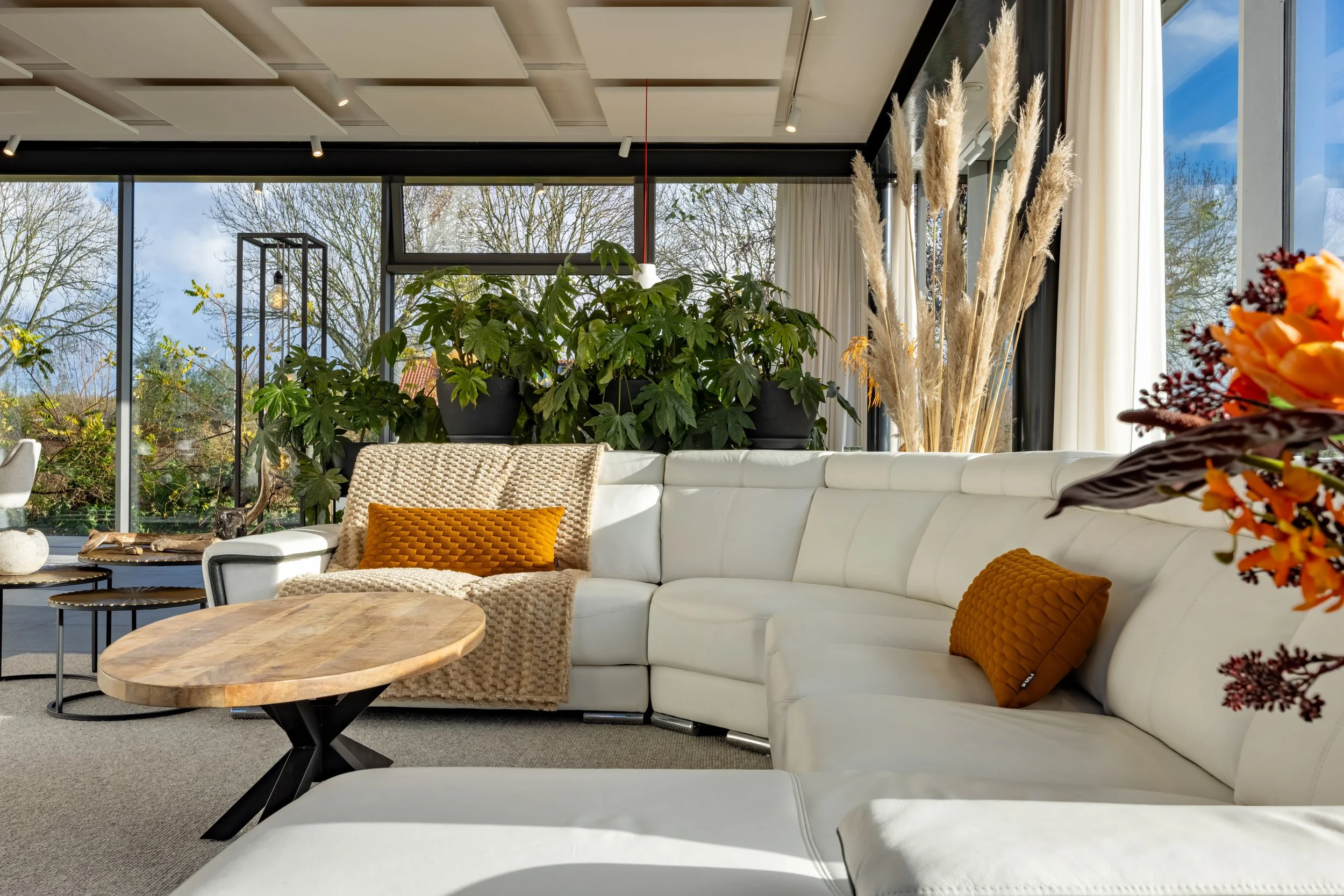 Licht woonkamer met grote witte hoekbank, oranjerode kussens, ronde houten salontafel, grote ramen, groene planten, decoratieve bloemen, en buitenzicht met bomen en blauwe lucht.