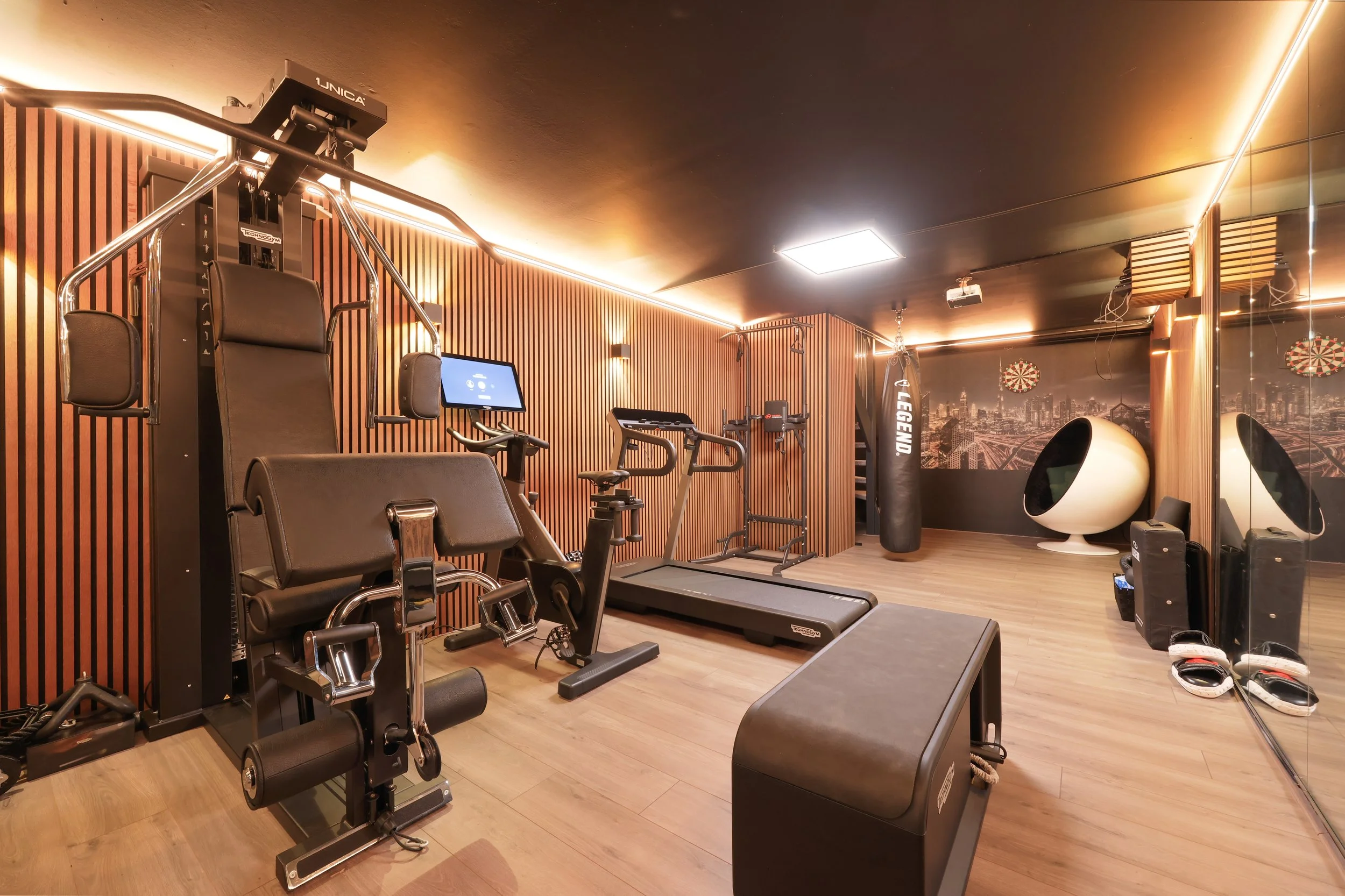 Klein modern thuisfitnessruimte met cardio-apparatuur, een gesponsorde bokszak, en een zitbal onder een muurschildering van een skyline, met glazen muur en houten vloer.