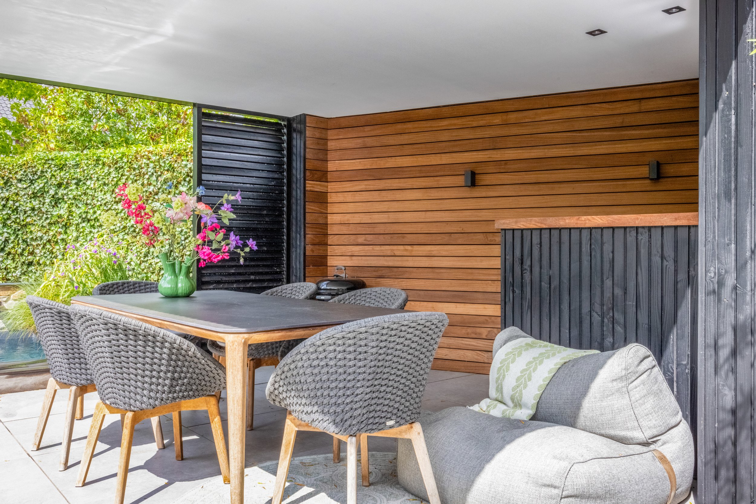 Terras met houten wand, zwart plafond, tafel met zes stoelen en een bank met kussen, groene vaas met bloemen, open schuifdeur naar tuin.