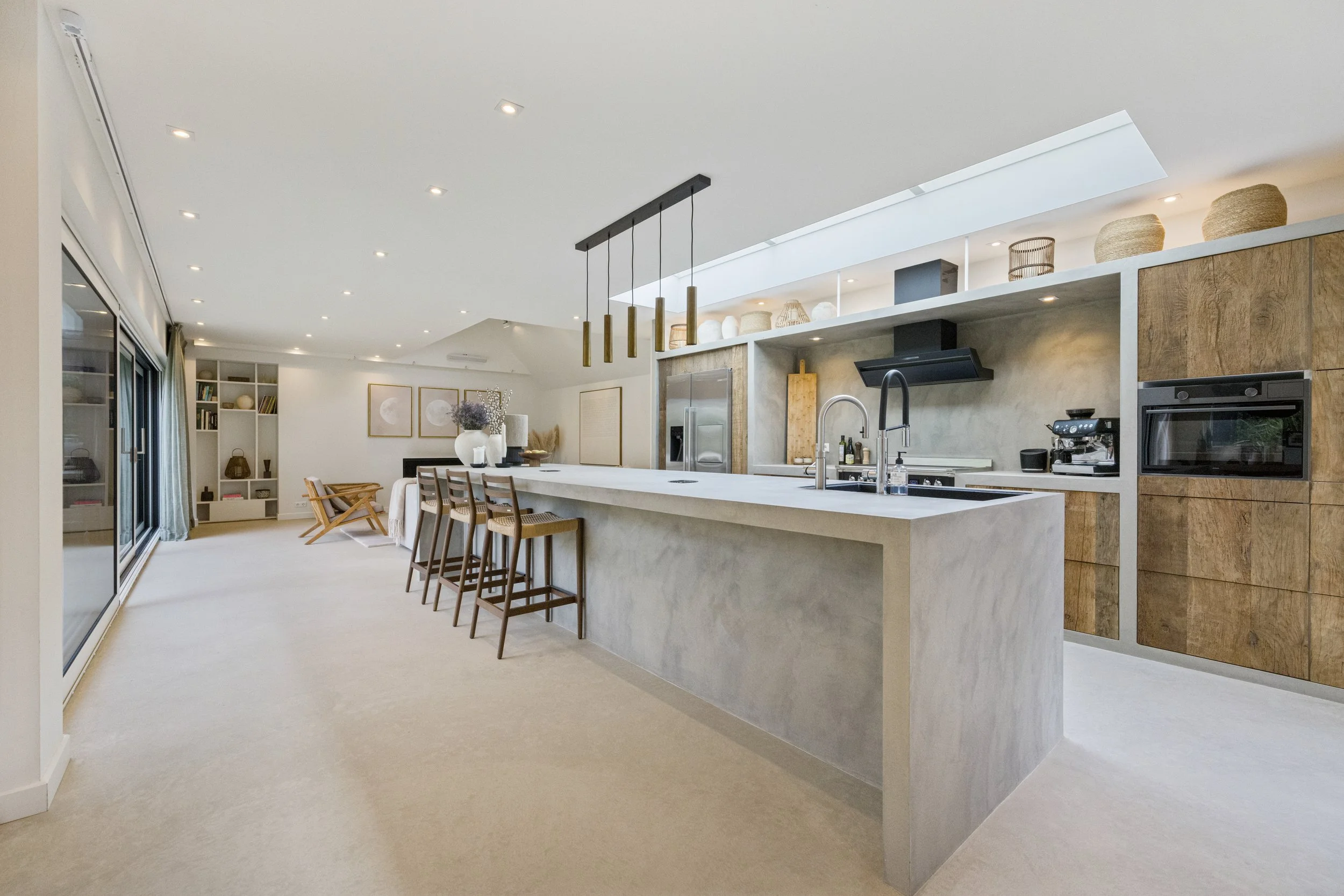 Een open keuken met een grote keukentafel en barkrukken, keukenkasten van hout, een fornuis, een magnetron en verschillende decoratieve elementen op open planken.