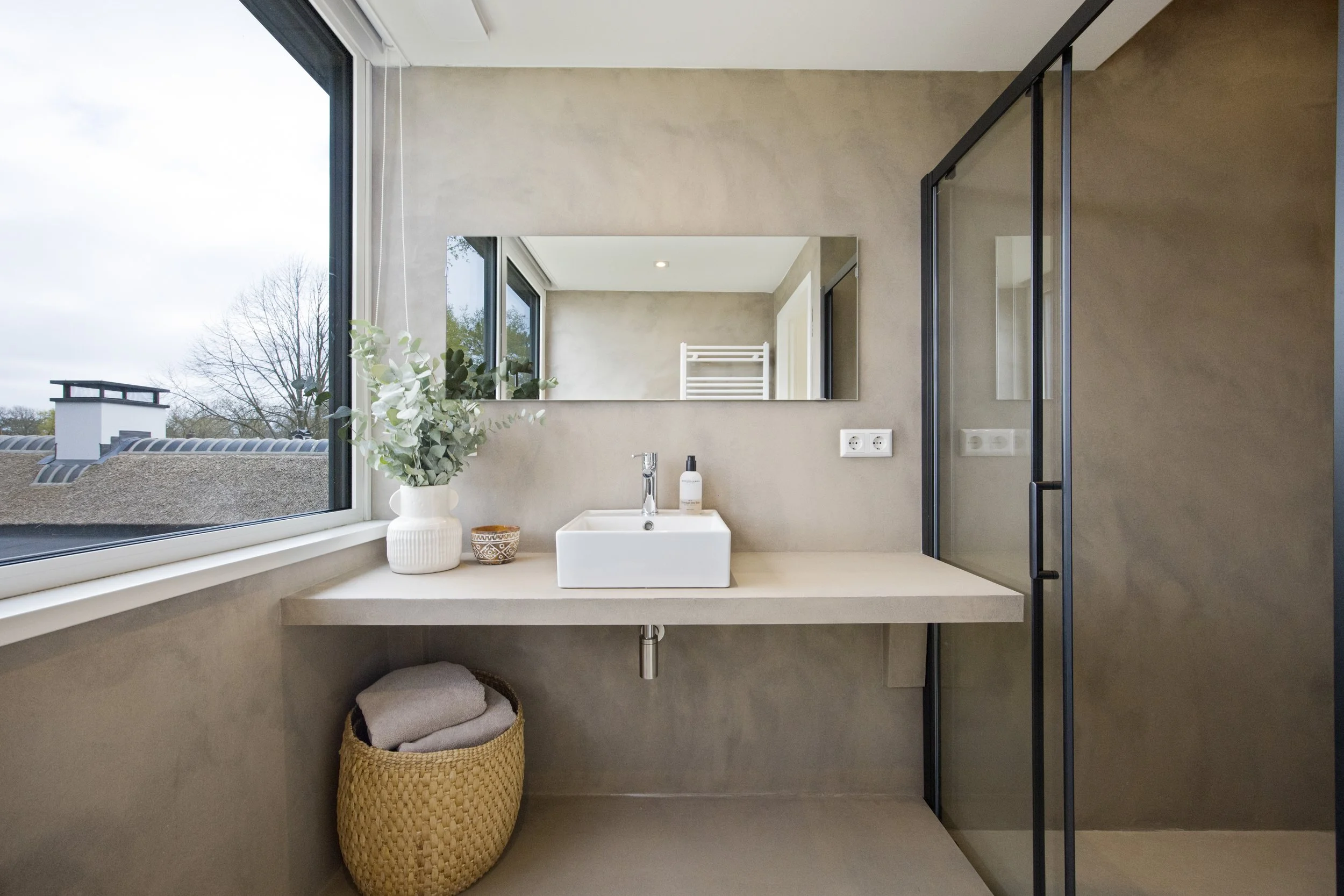 Moderne badkamer met groot raam, witte wastafel, spiegel, glasdouche en een rieten mand met handdoeken.