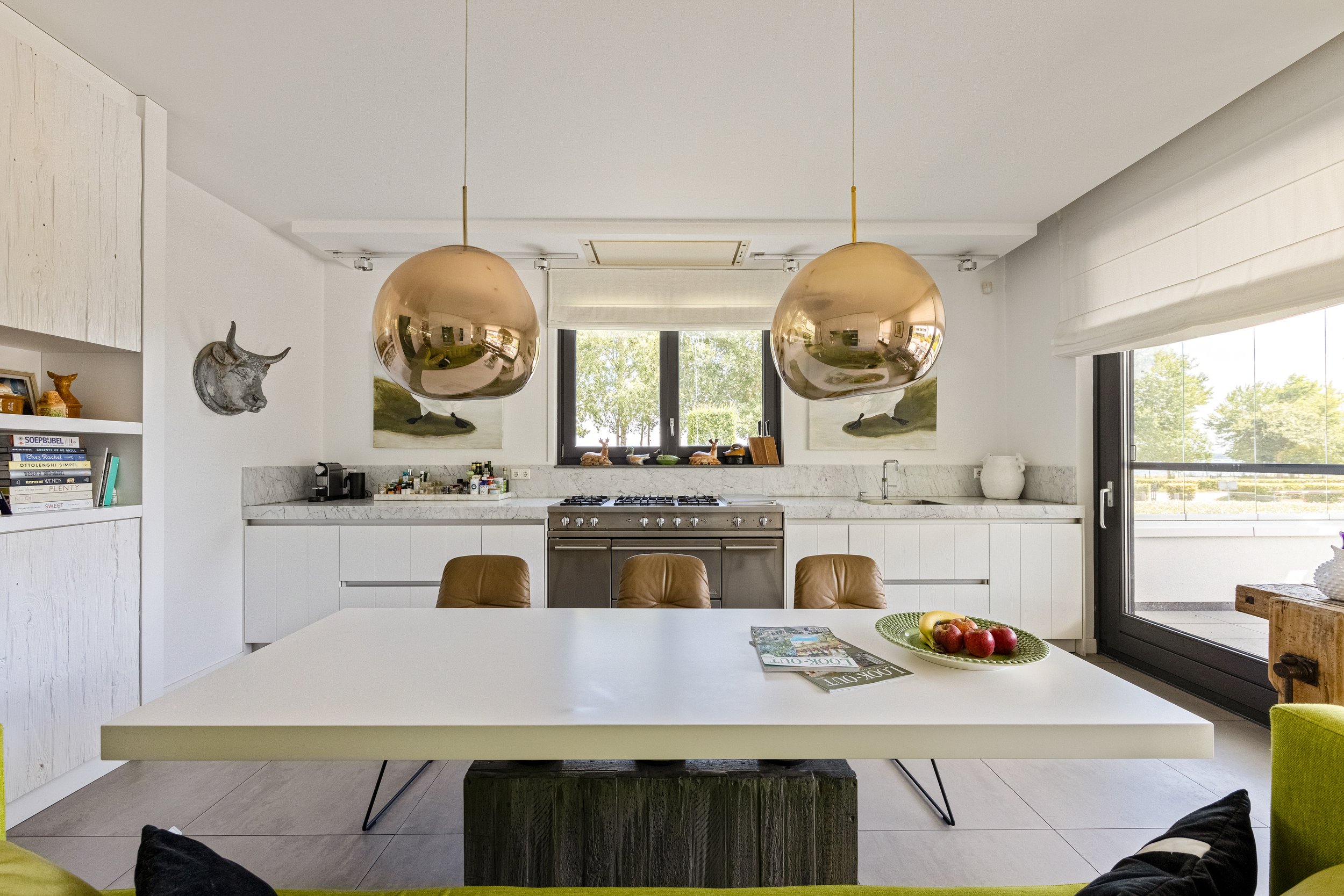 Moderne keuken met witte kasten, een gasfornuis, twee grote gouden hanglampen, een raam boven het fornuis en een deur naar het balkon, met een witte tafel en bruin leren stoelen.