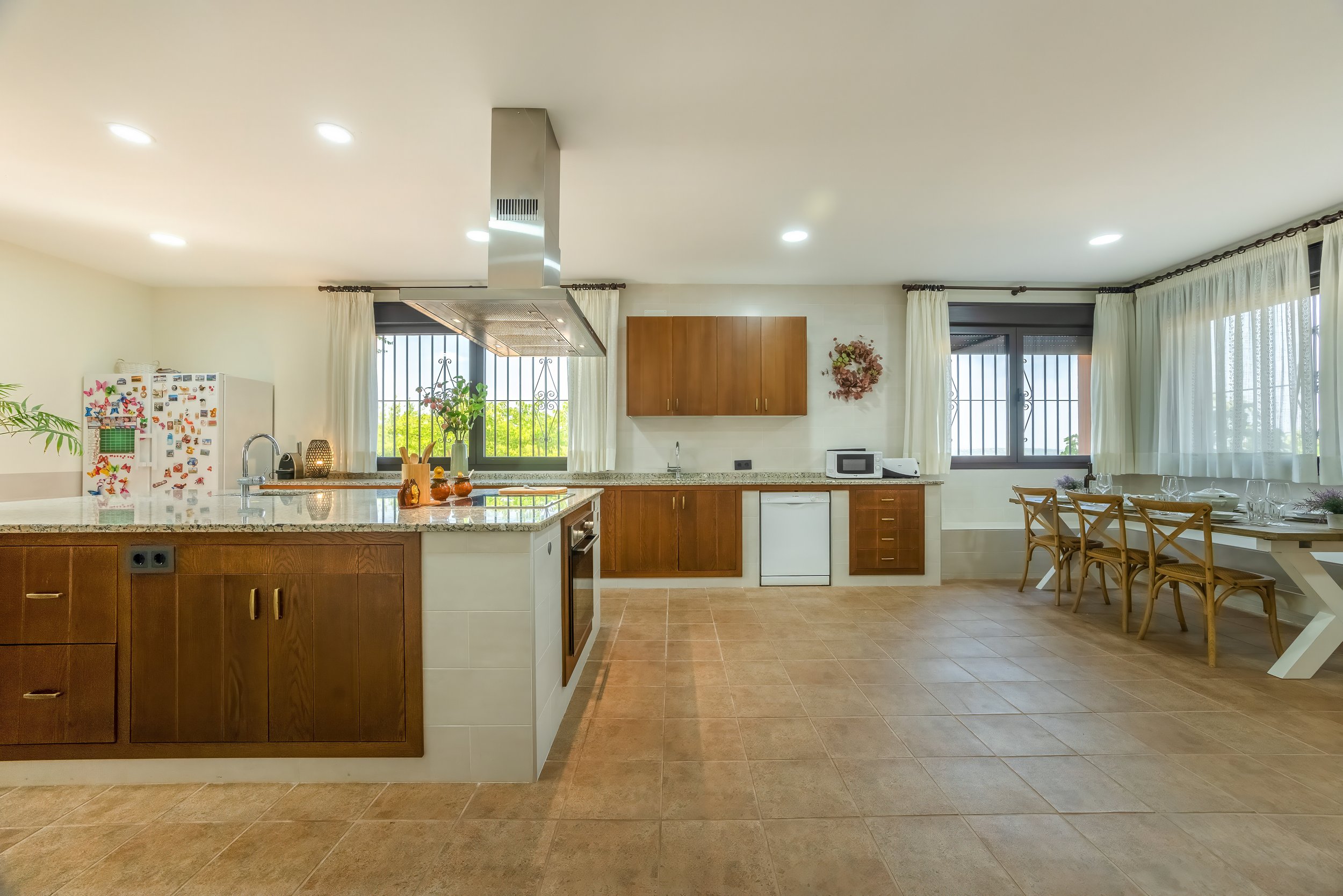Grote keuken met houten kasten, een kookeiland en een eetgedeelte met houten stoelen voor een raam, veel natuurlijk licht en witte gordijnen.