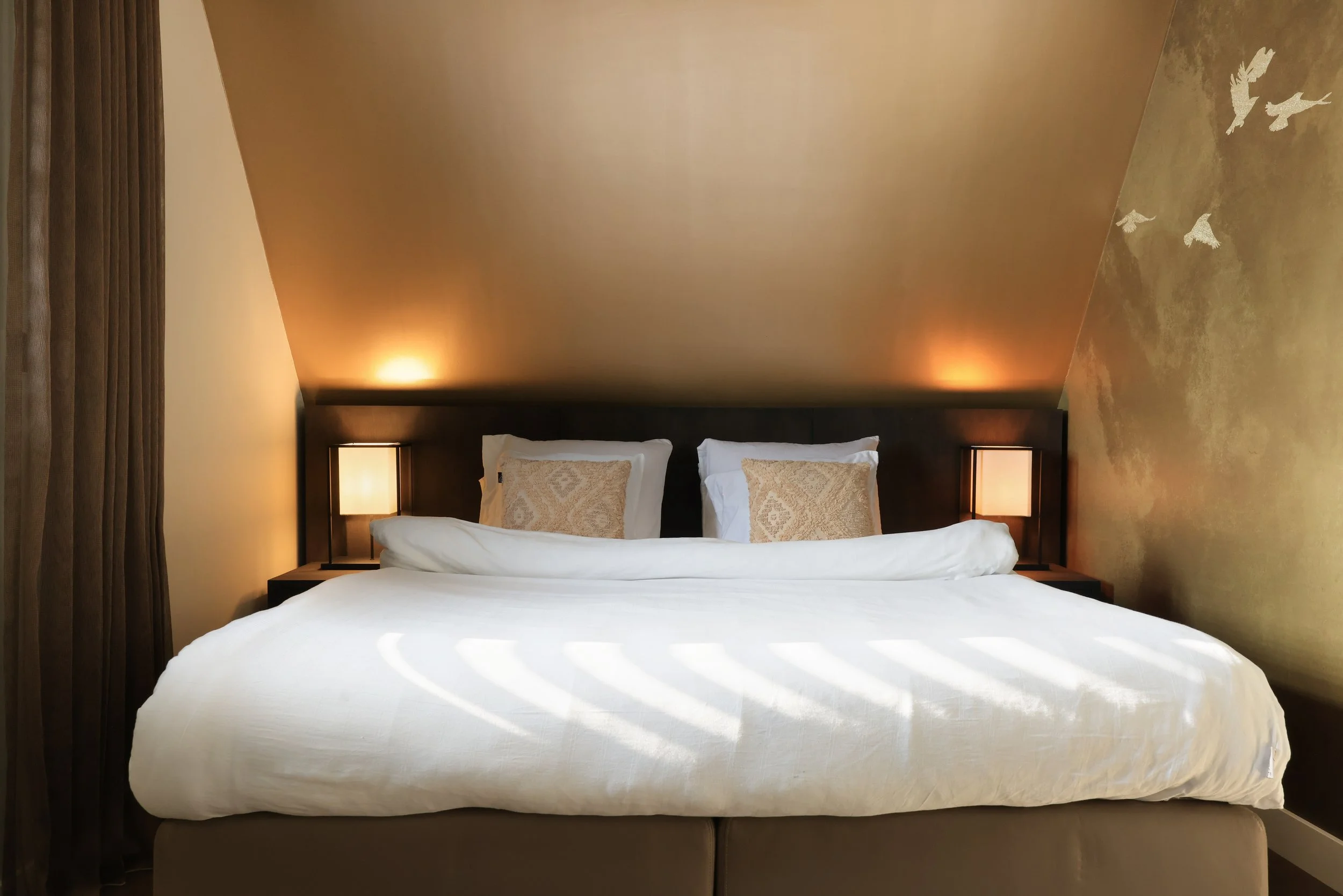 Een bed met wit beddengoed en twee kussen, zachte verlichting aan beide kanten, een muur met een muurschildering van vogels.
