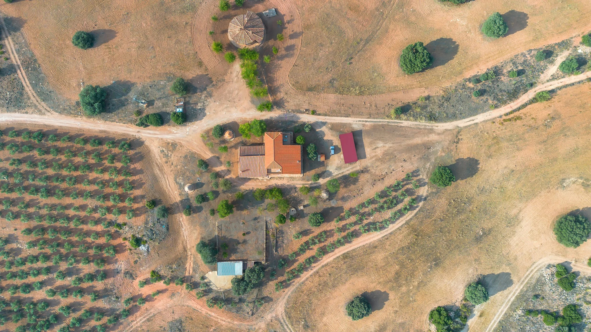 Een vogelvluchtfoto van een landelijk gebied met een huis met rood dak, omgeven door bomen, tuinen, en velden, met aarde en paden.