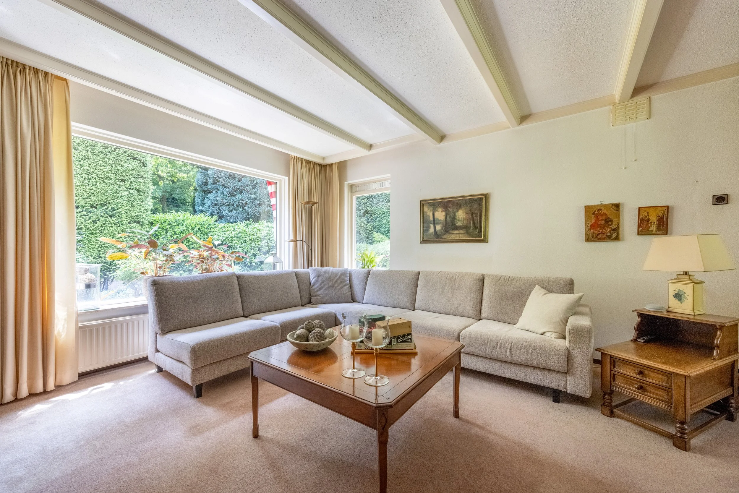 Woonkamer met grote hoekbank, houten salontafel, schilderijen aan de muur, grote ramen met uitzicht op een tuin en zonlicht.