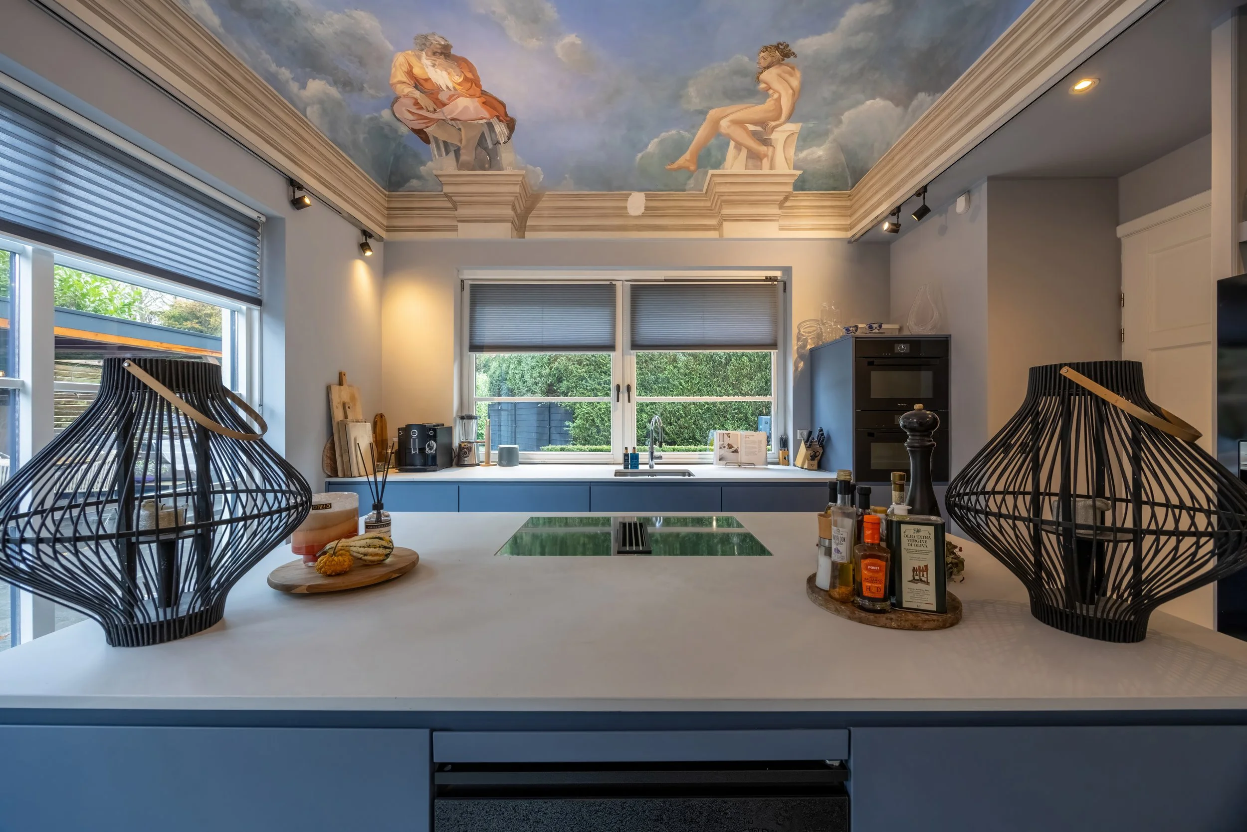 Moderne keuken met wit eiland, twee zwarte kandelaars, keukenapparatuur, ramen met uitzicht op tuin, plafondschildering met twee oude filosofen op de wolken.