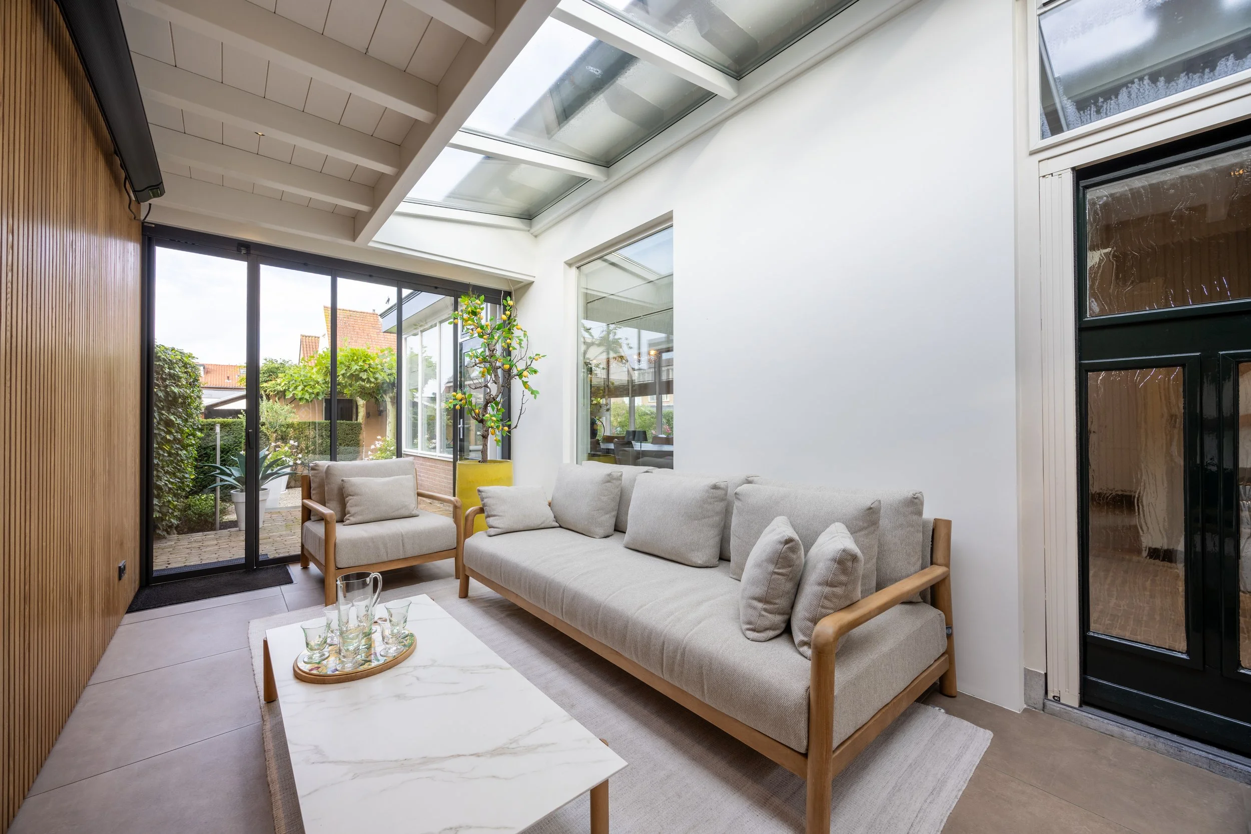 Een lichte woonkamer met een grote loungebank, een salontafel met glazen en een houten kast, veel grote ramen en een glazen dak, en uitzicht op een tuin.