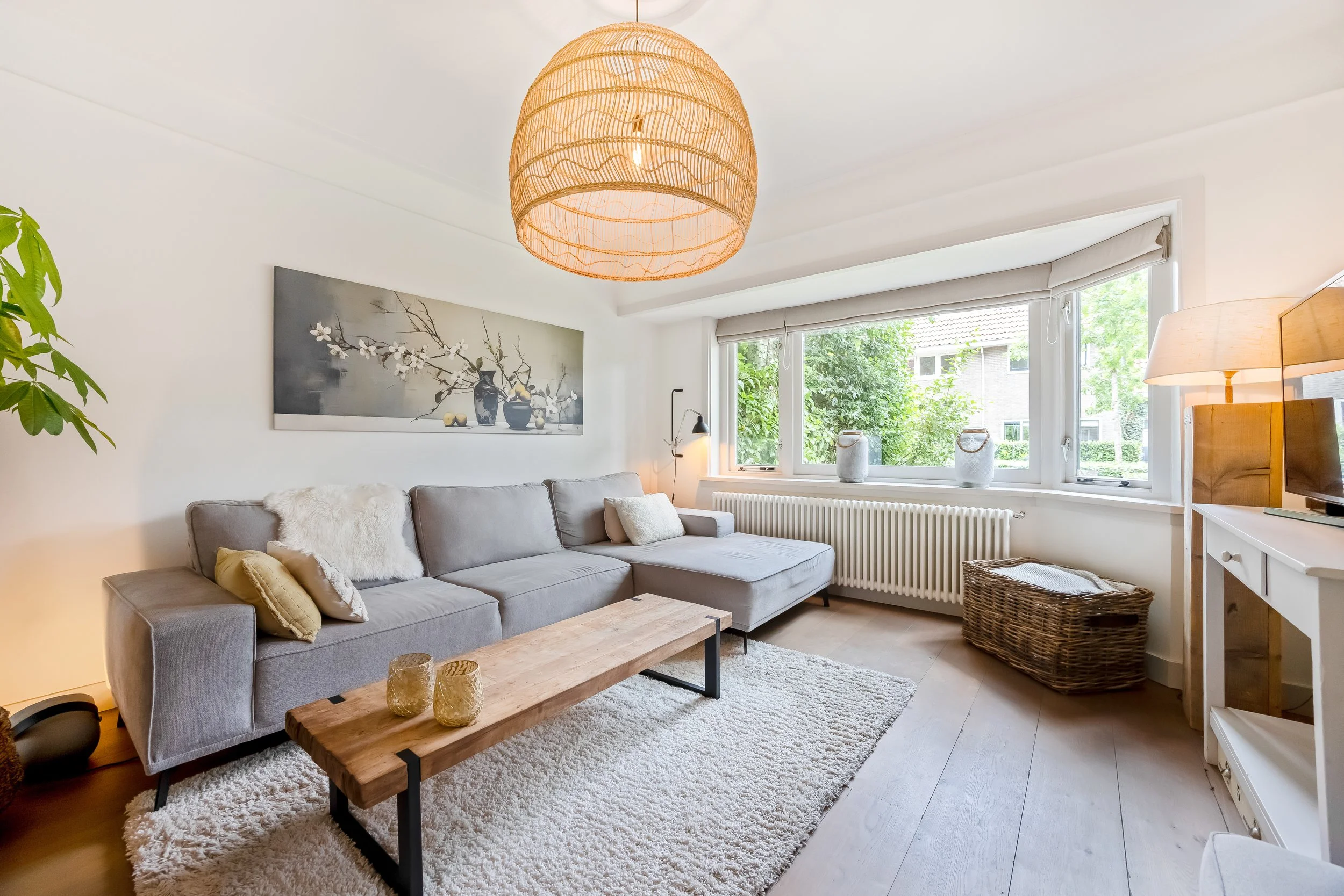 Lichte woonkamer met groot raam, grijze hoekbank, houten salontafel, vloerkleed, muurkunst, planten, en decoratieve objecten.