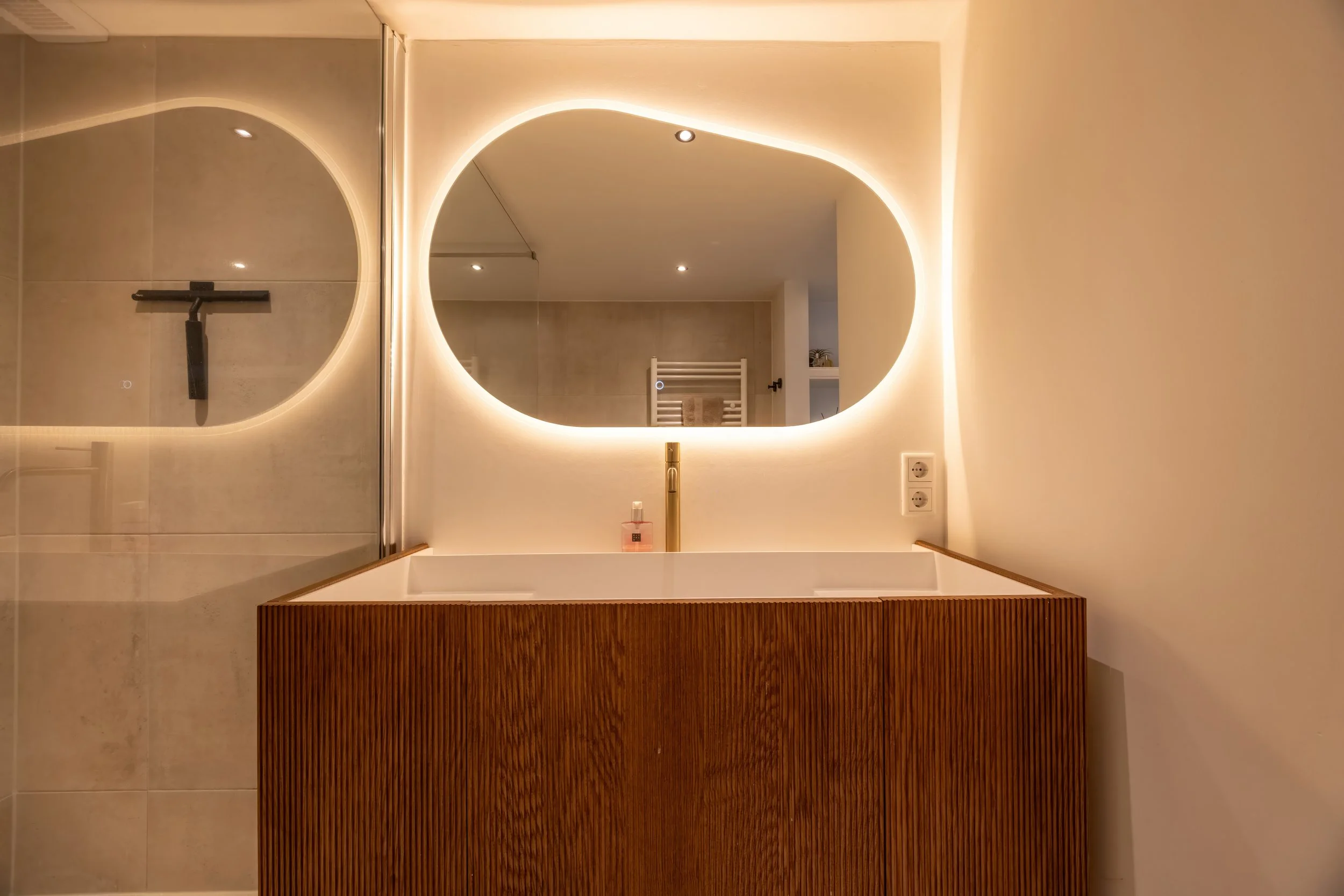 Moderne badkamer met een lichtgekleurde wand en een ronde spiegel met ledverlichting, een houten wastafel en een gouden kraan.