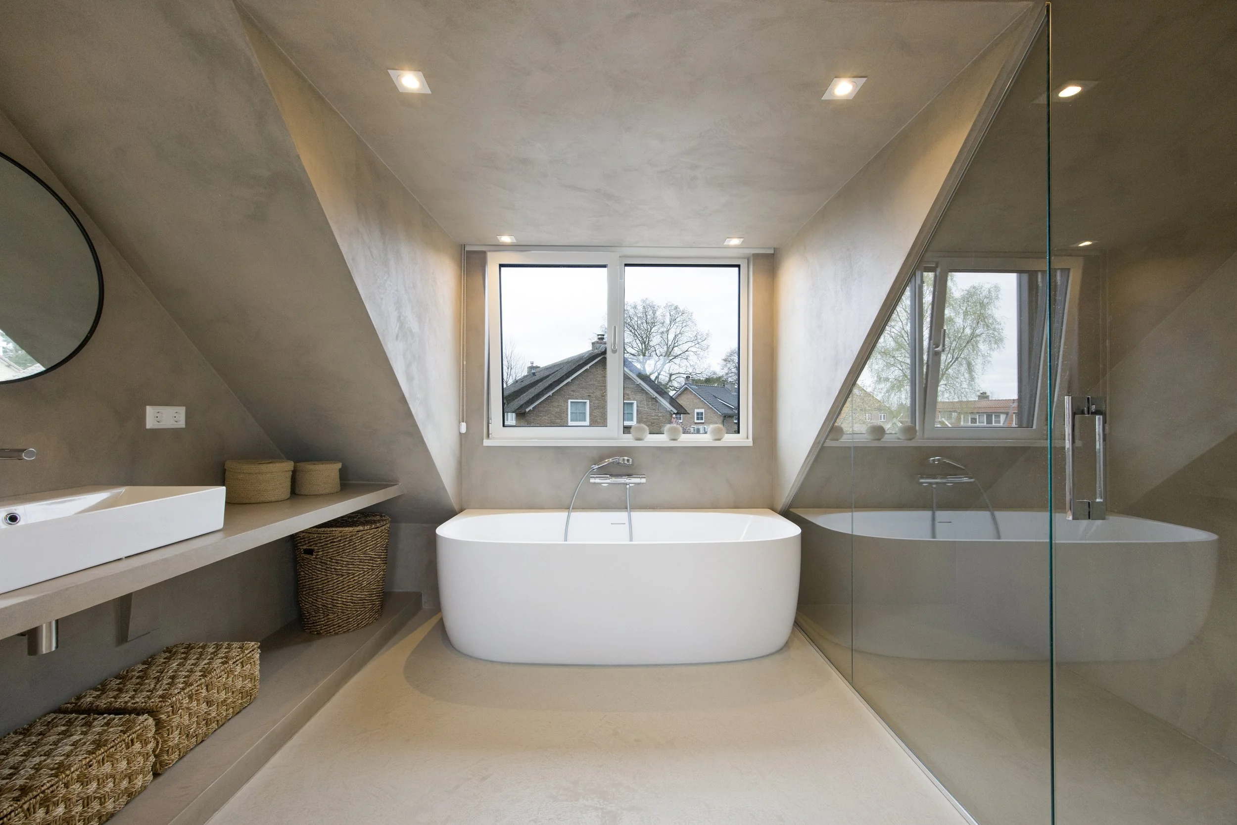 Moderne badkamer met vrijstaande badkuip onder groot raam, jacuzzi met glazen wand, wastafel met ronde spiegel, en Romeinse-poedermuur, beige vloer en plafond