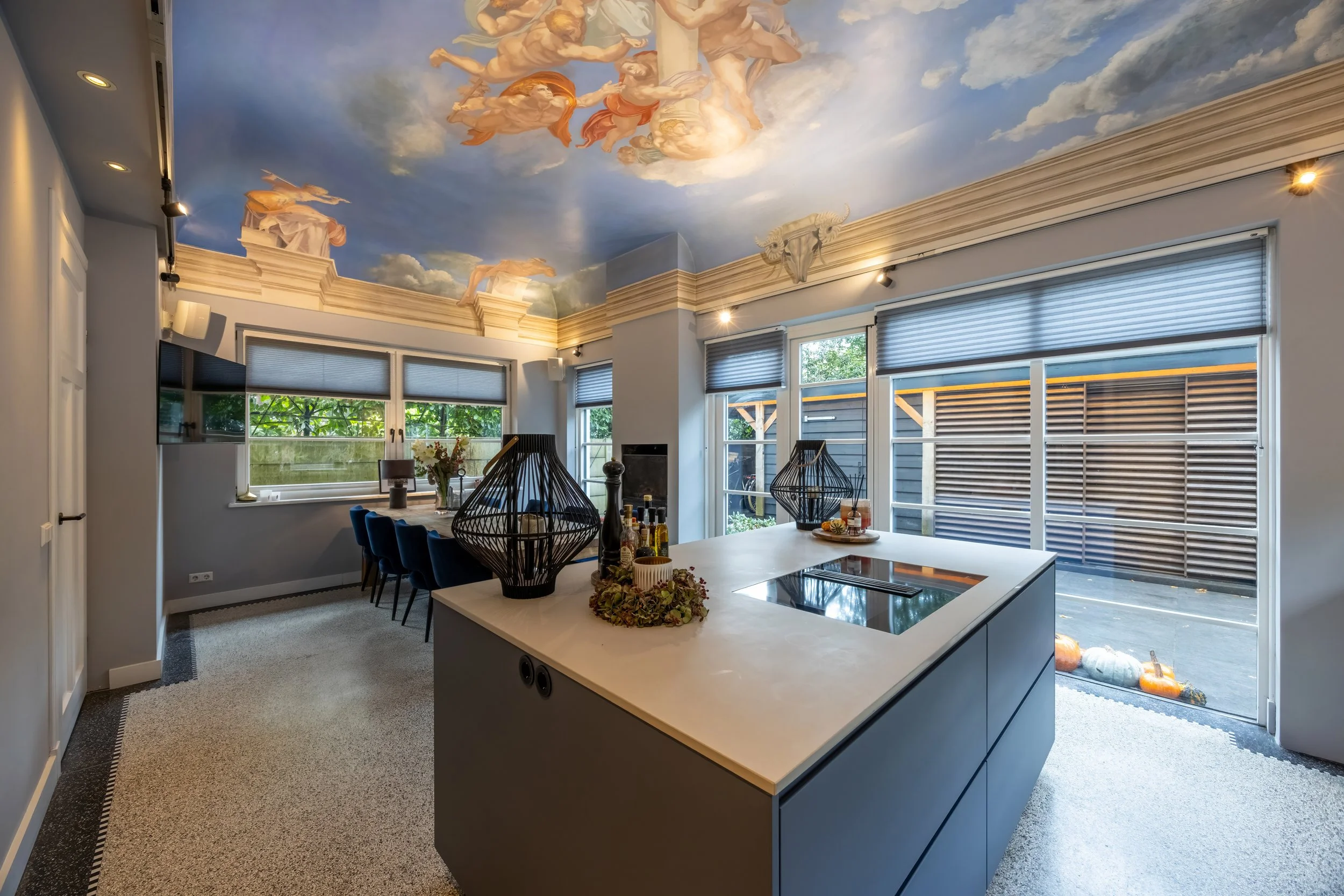 Een keuken met een eiland en een eettafel, een knalblauwe plafondschildering van goddelijke figuren, grote ramen met blauwe jaloezieën, een open tuin met tuinbeelden en pompoenen buiten.