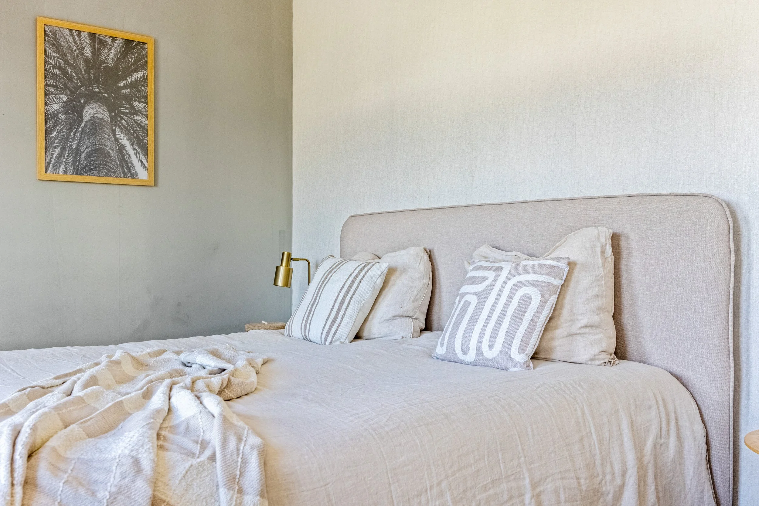 Een comfortabel slaapkamer met een beige bed met meerdere kussens en een lichtgekleurde deken, naast een nachtkastje met een gouden leeslamp.