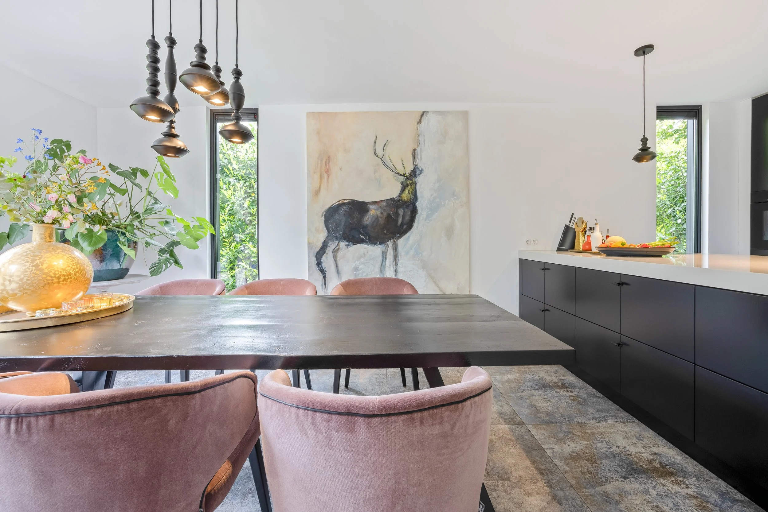 Een moderne eetkamer met een grote houten tafel, roze velvet stoelen, een grote gouden vaas met bloemen, een schilderij van een hert op de muur, en zwarte keuken kasten.