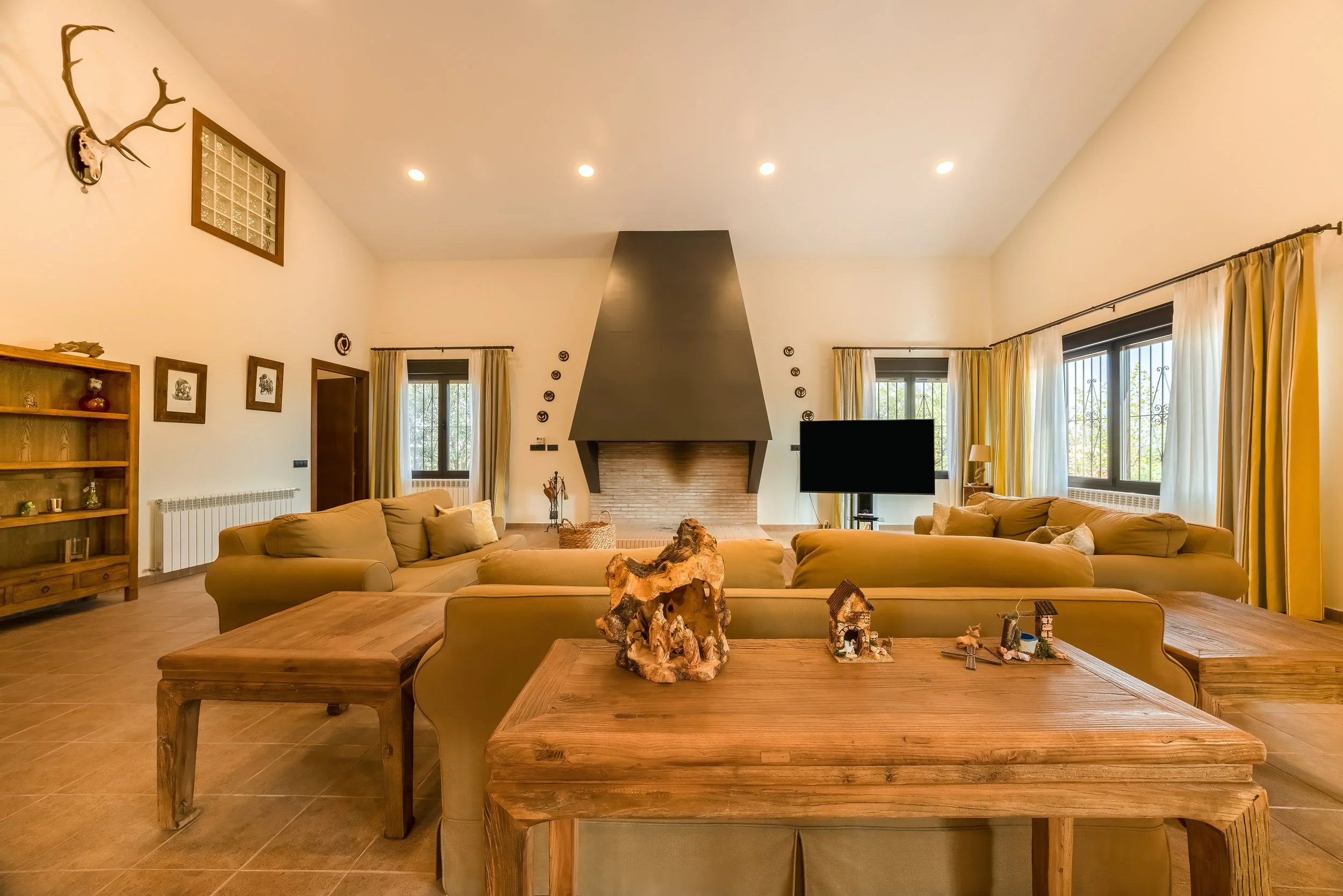 Gezellige woonkamer met grote ramen, beige banken, een zwarte schoorsteen, houten meubels en decoraties, en natuurlijke lichtinval.